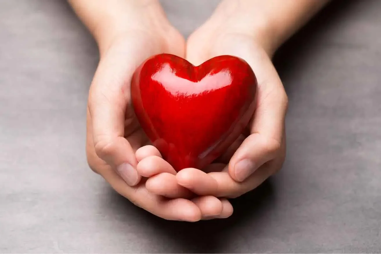 Person holding a shiny red heart-shaped object with both hands, over a blurred gray background.