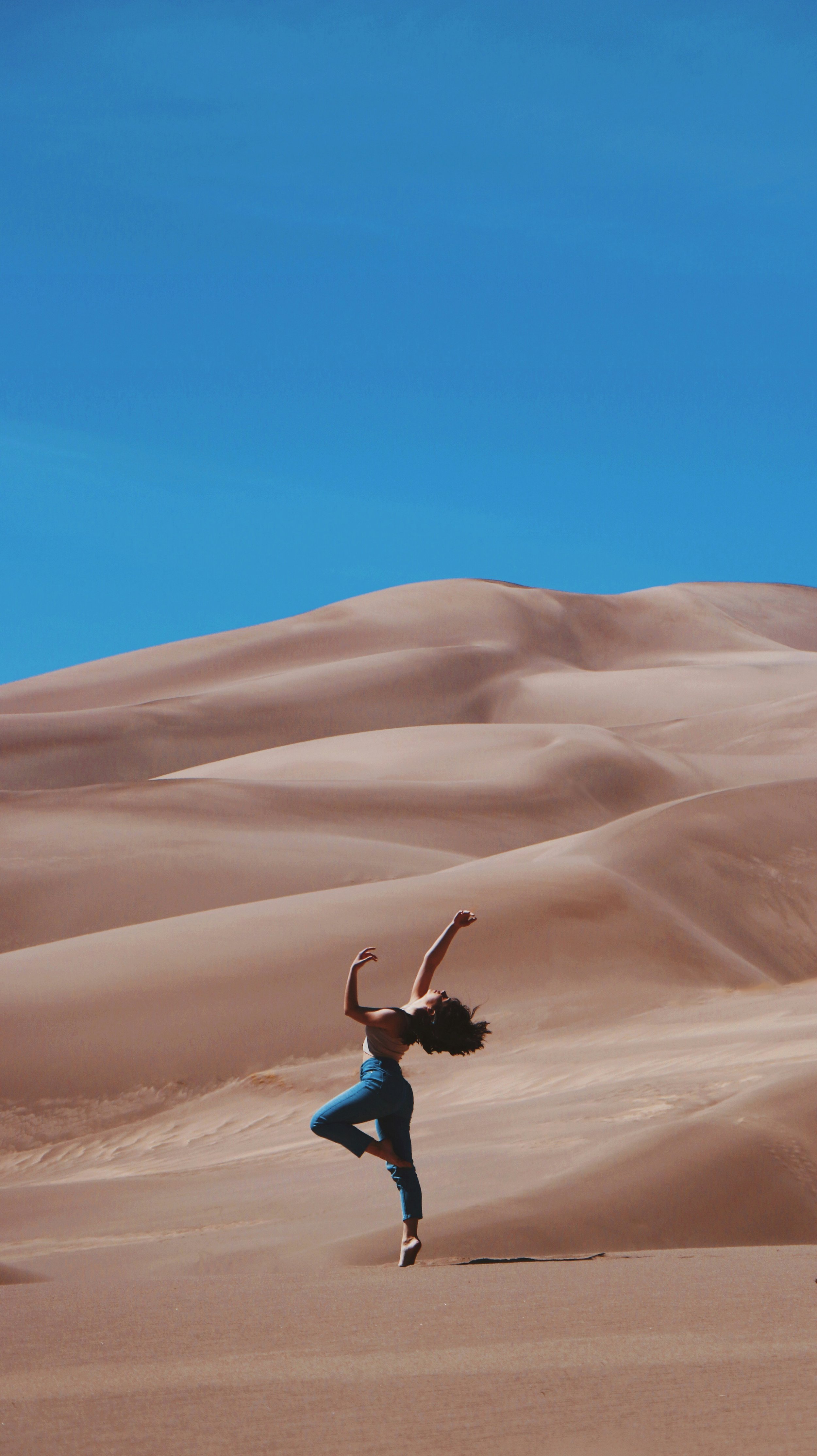 Person in blue pants and a black top dancing on sand dunes under a clear blue sky.