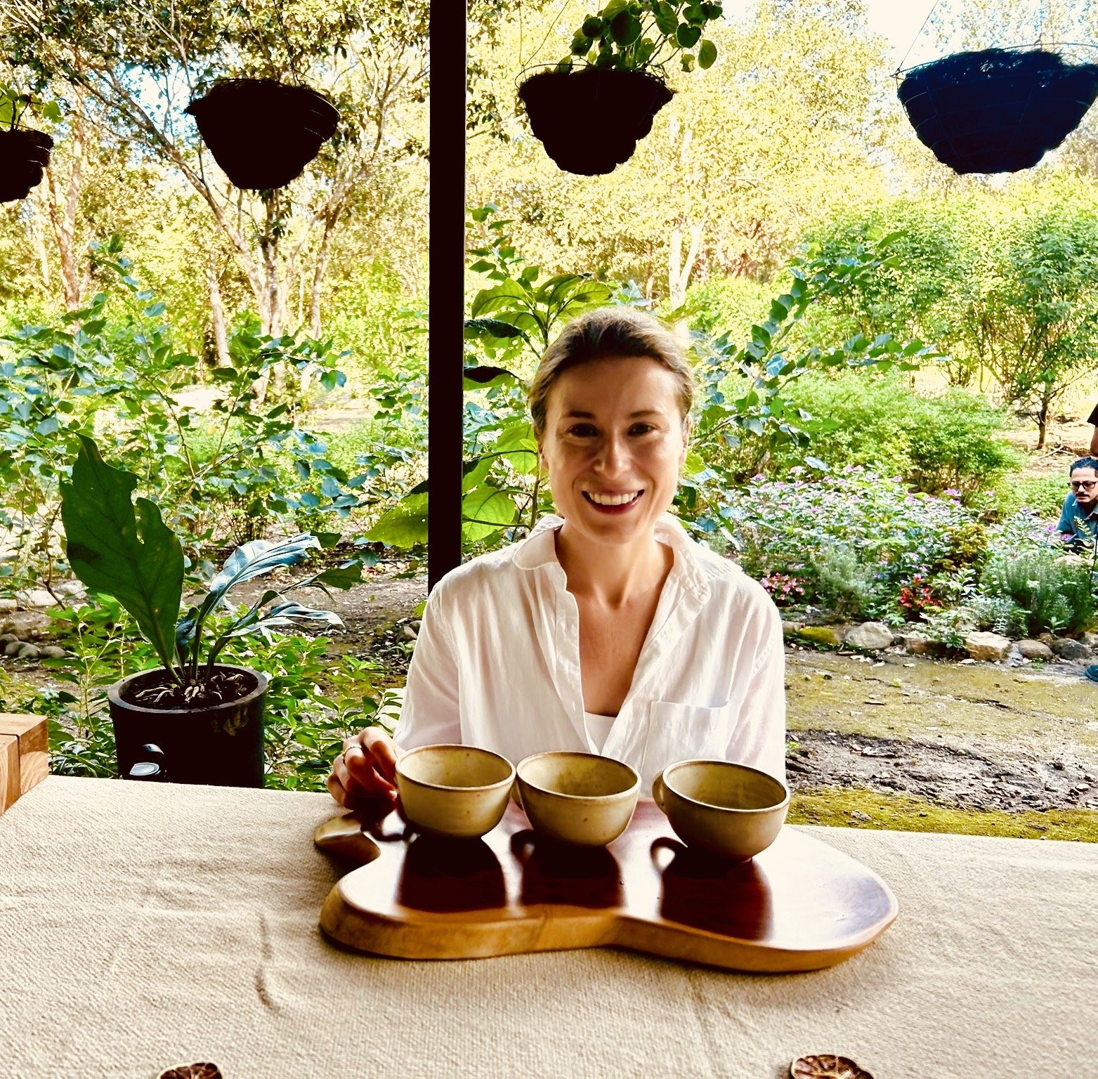 Person sitting at a table with three ceramic cups on a wooden tray in a garden setting, surrounded by green plants and hanging pots.