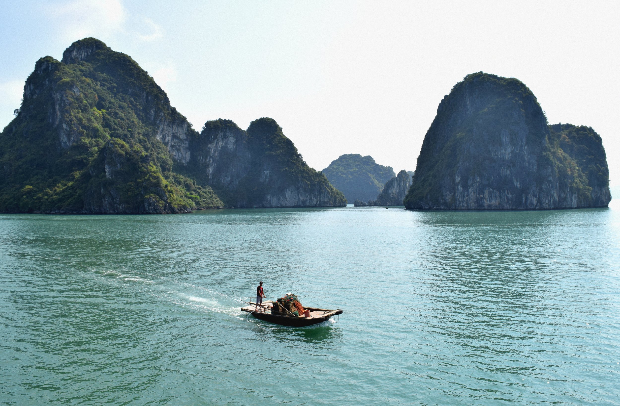 A boat with a person on it floating on turquoise water near towering green-covered limestone islands in the distance under a partly cloudy sky.