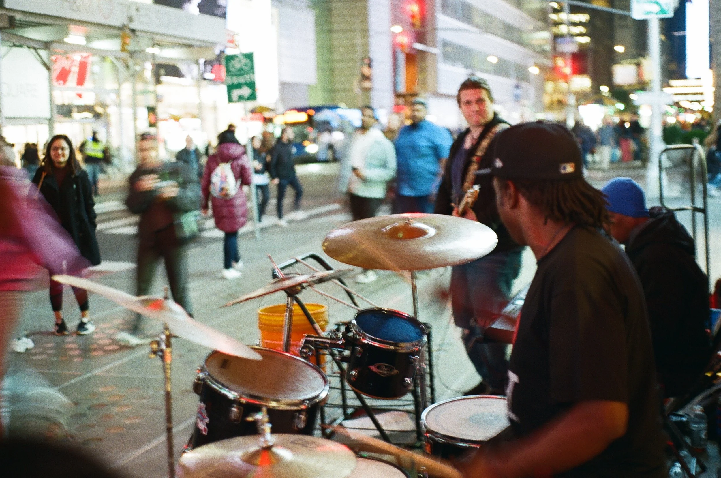 Street musician playing drums in a busy city street at night, with pedestrians walking by and illuminated storefronts in the background.