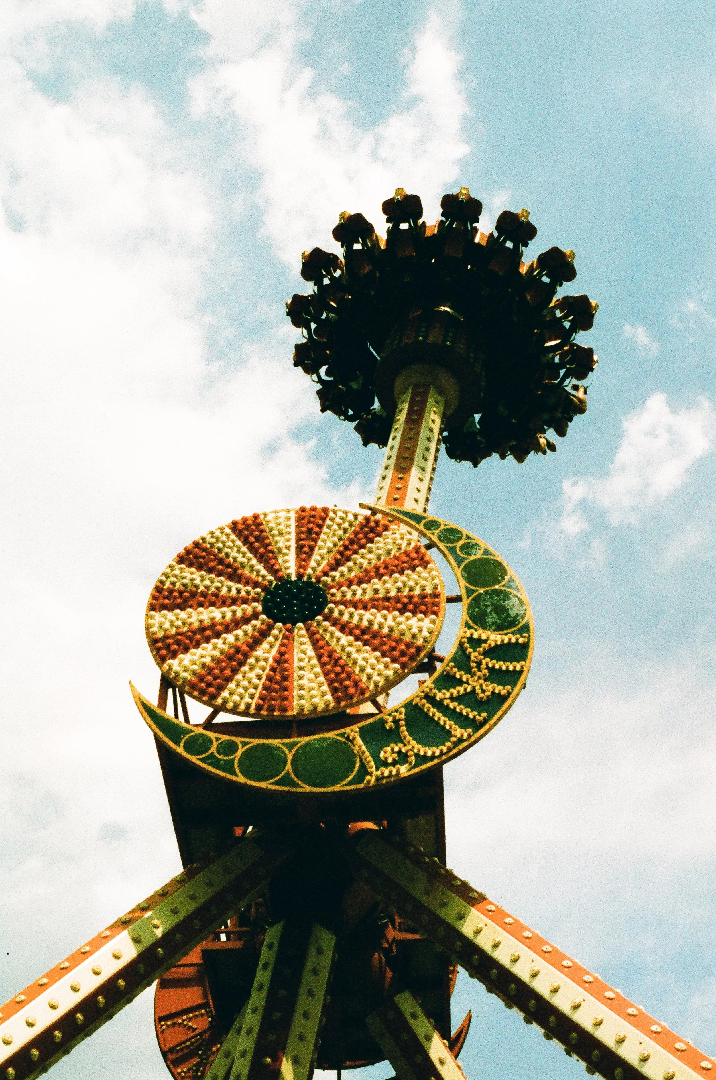 A tall amusement park ride featuring a circular spinning component at the bottom with lights and designs, and a head of seats at the top with passengers, set against a partly cloudy sky.