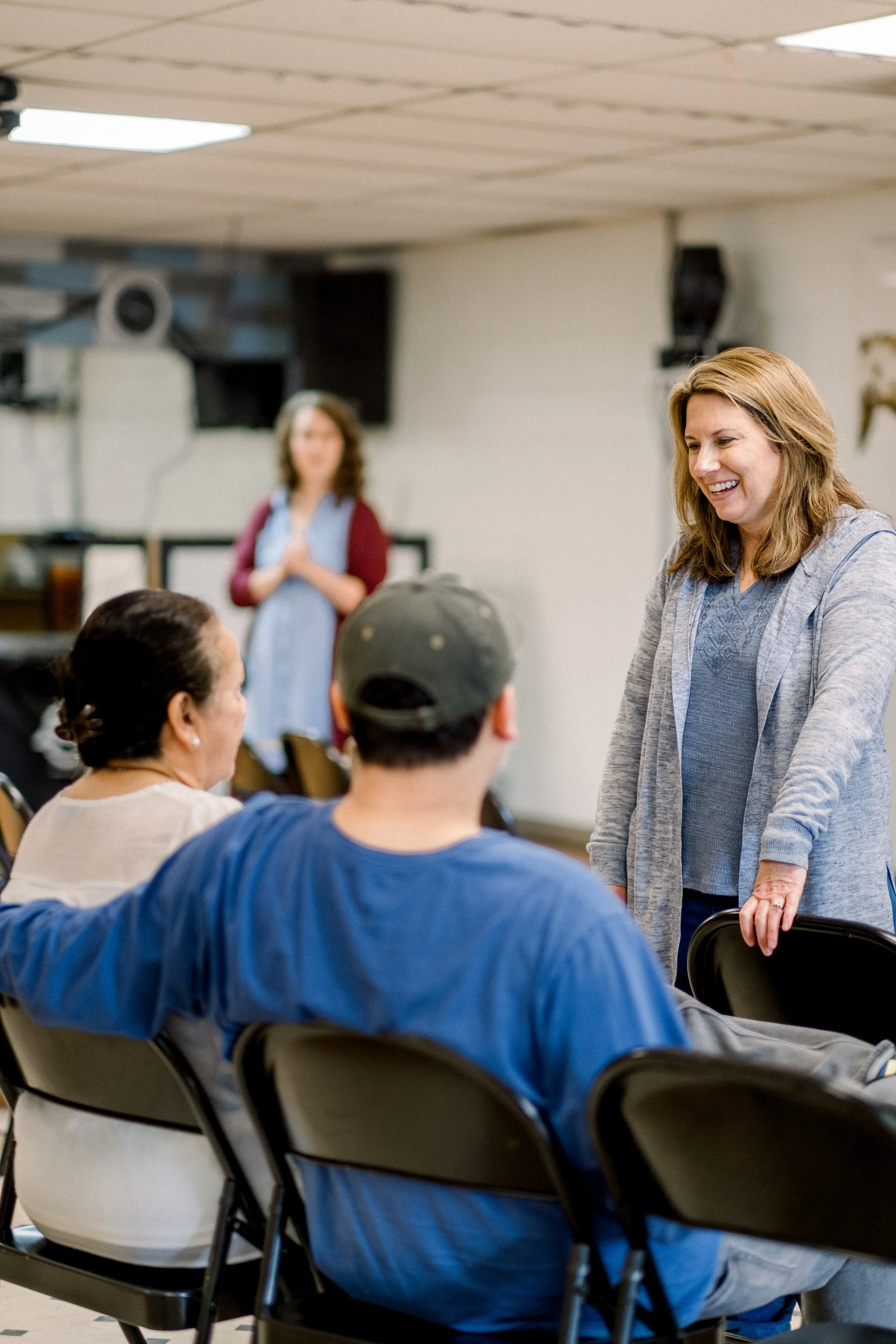 A woman with shoulder-length hair smiling and talking to a group of people seated in a room, with another woman standing in the background.