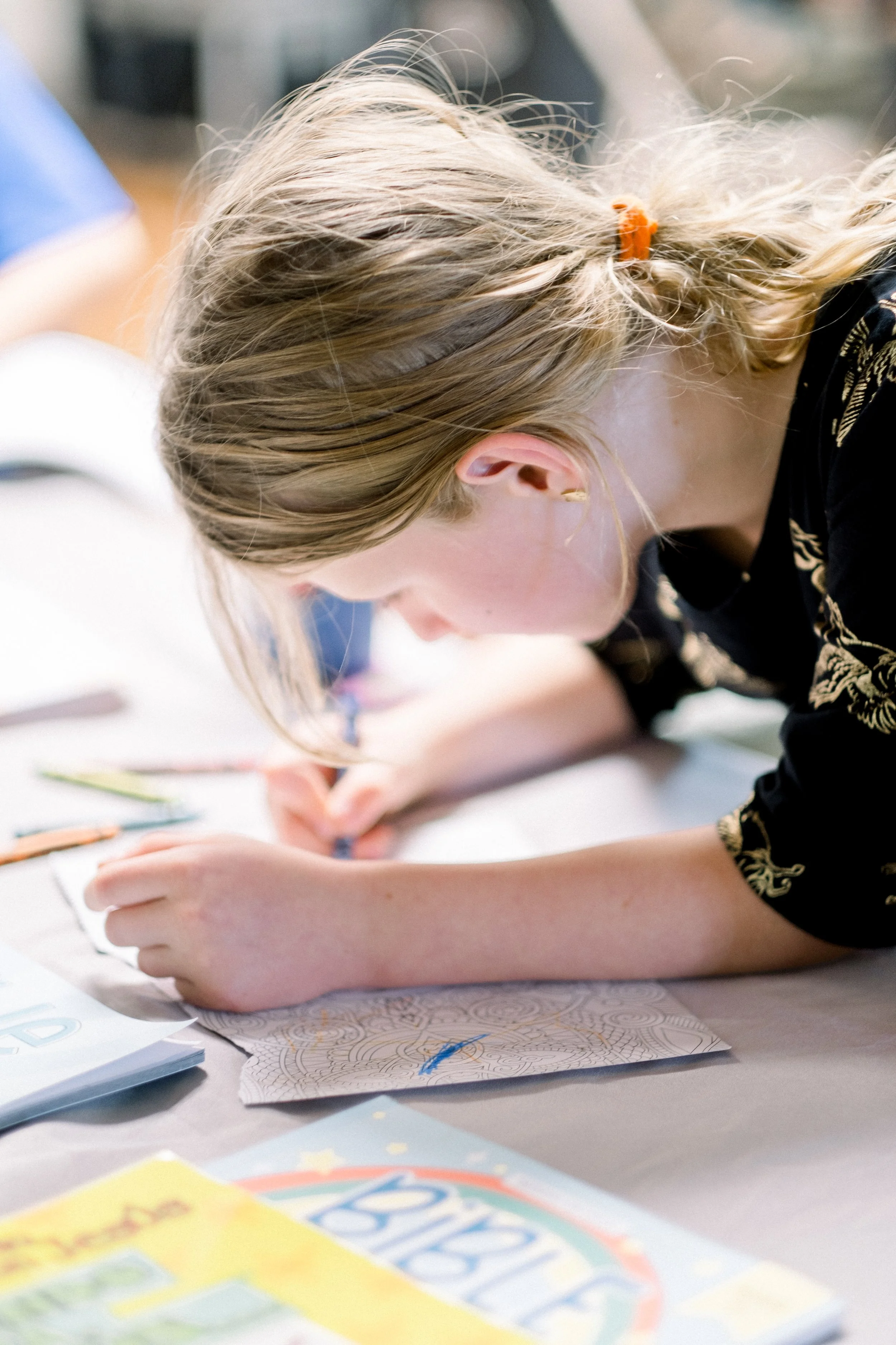 A young girl with long blonde hair tied back with an orange hair clip is focused on drawing on a piece of paper on a table, surrounded by colorful art supplies and a book.
