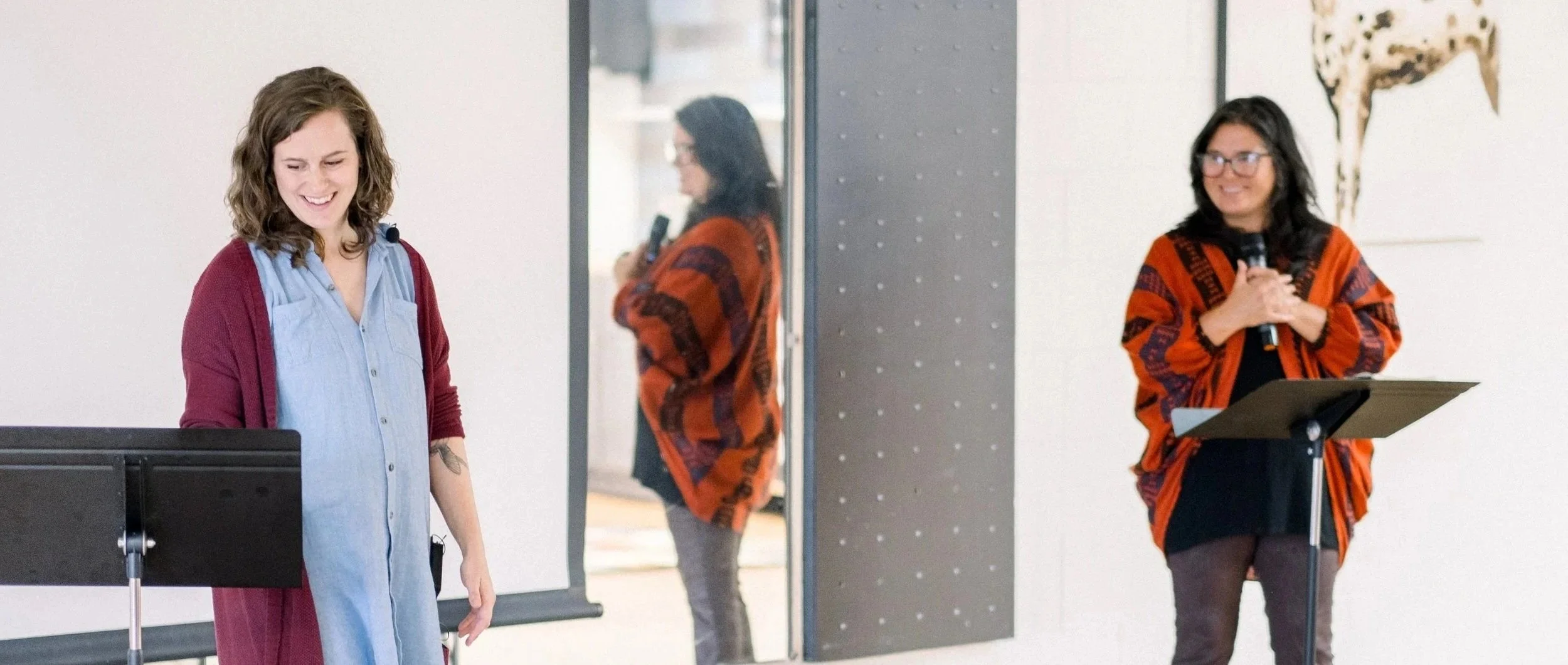Two women standing in a room, one is smiling and looking down, the other is holding a microphone and standing behind a music stand.