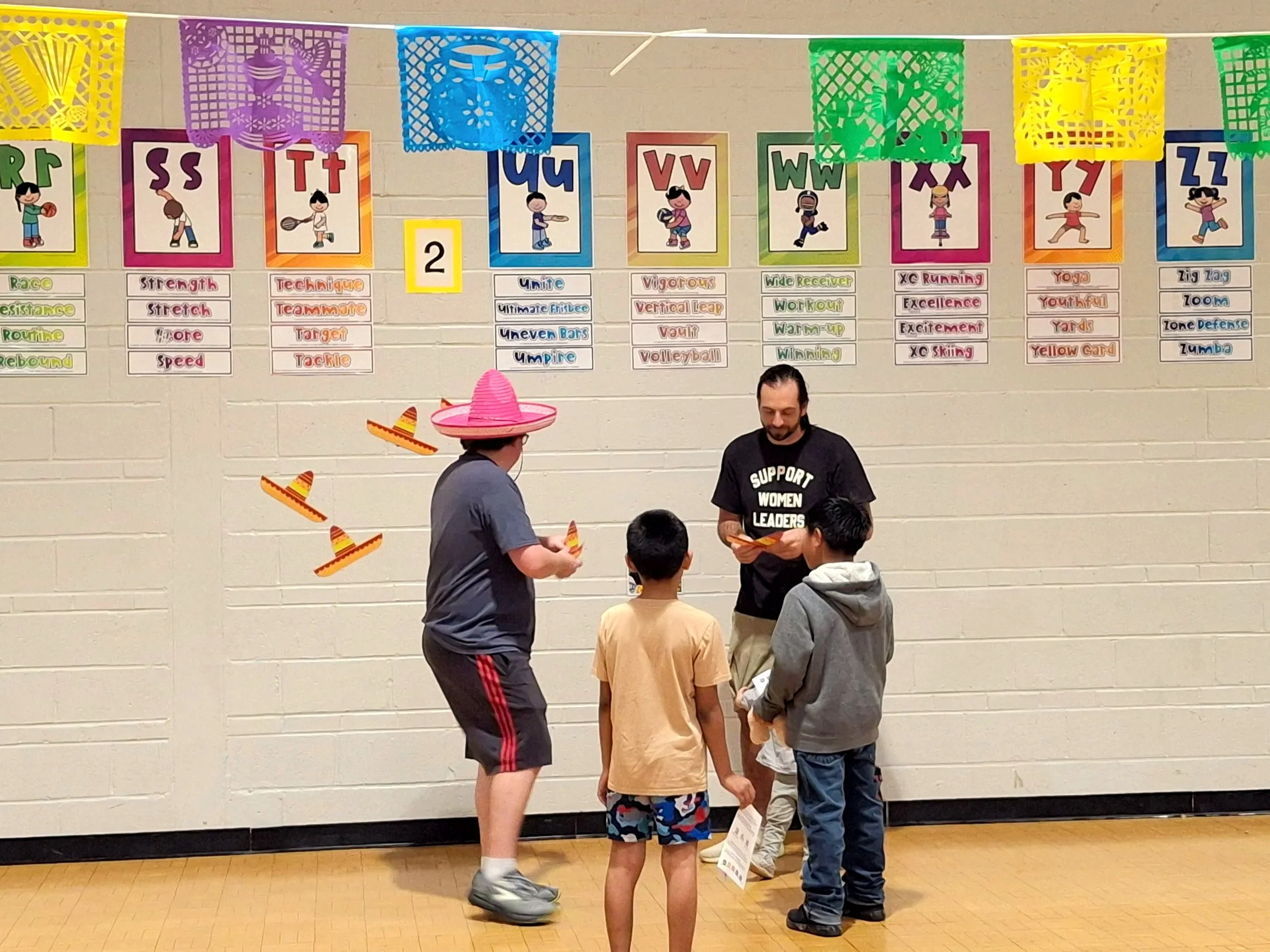 Adults and children standing in a gymnasium, with colorful posters and banners hanging on a wall behind them. One adult is wearing a pink sombrero, and there are orange magic hat stickers nearby, while the wall displays posters related to sports and activities.