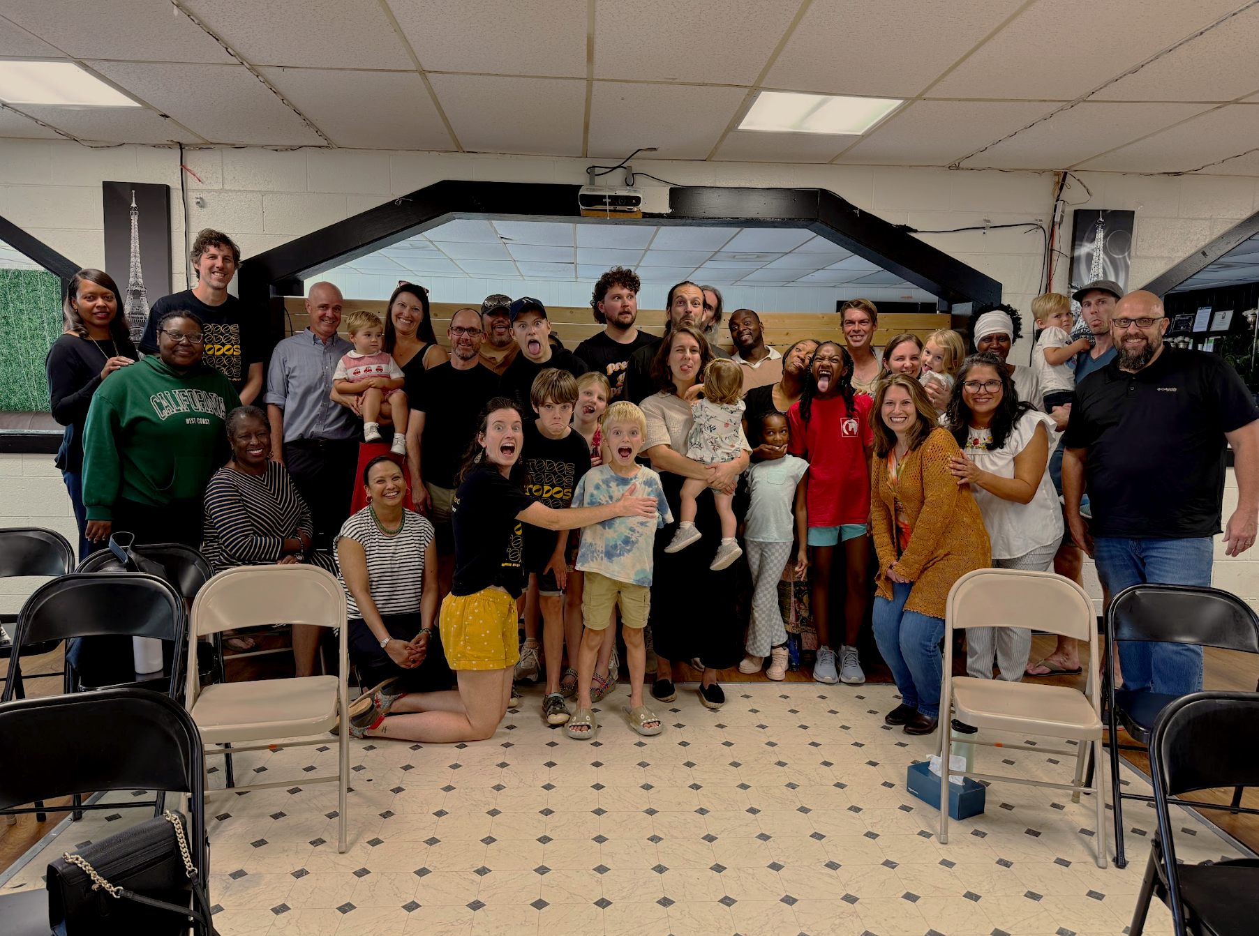 A large group of people of various ages and ethnicities gathered in a room for a photo, some seated, some standing, with a festive and joyful atmosphere.