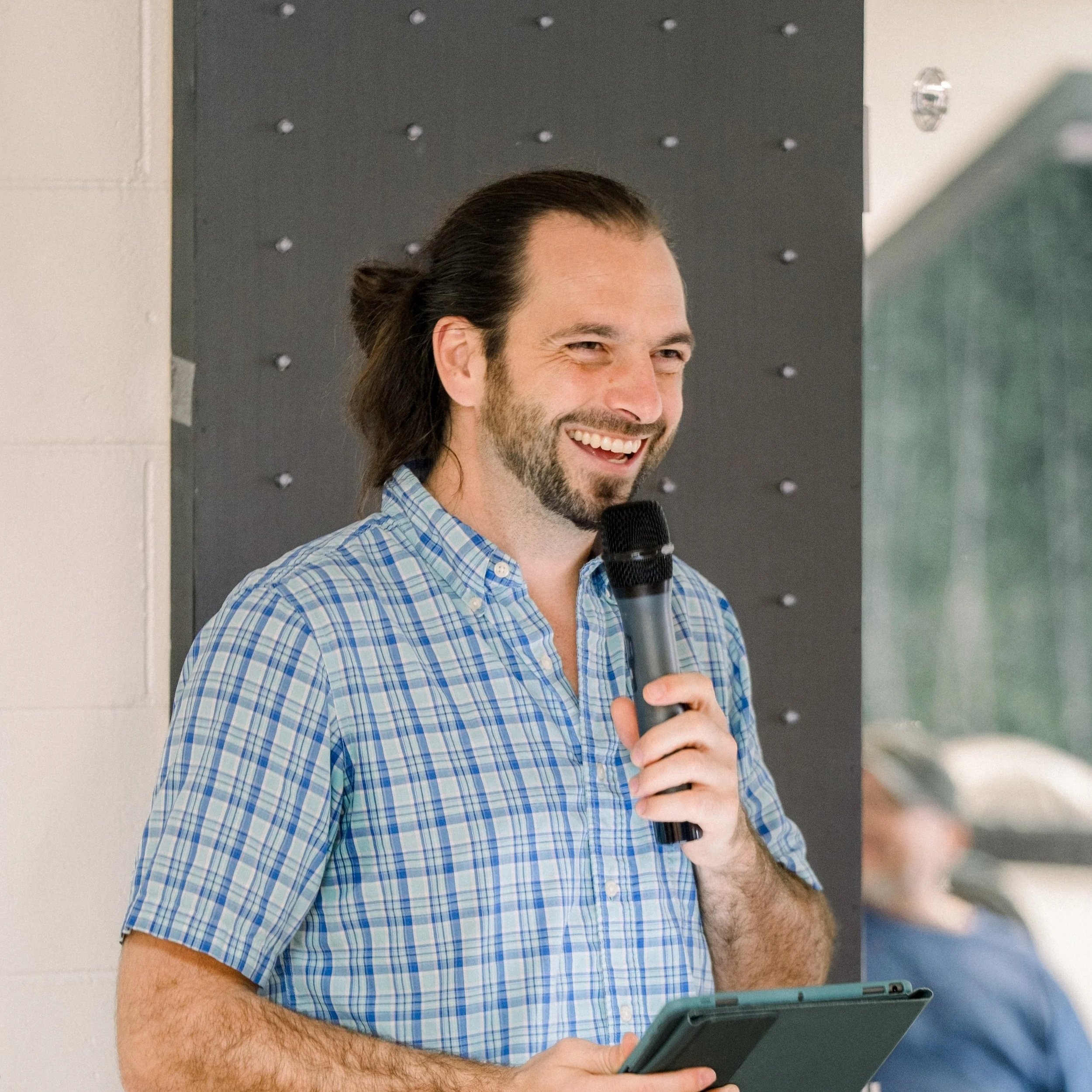 A man with long hair and a beard, wearing a blue checked shirt, smiling and holding a microphone and a tablet, standing in front of a blackboard or wall with a background that includes a window and a person sitting.