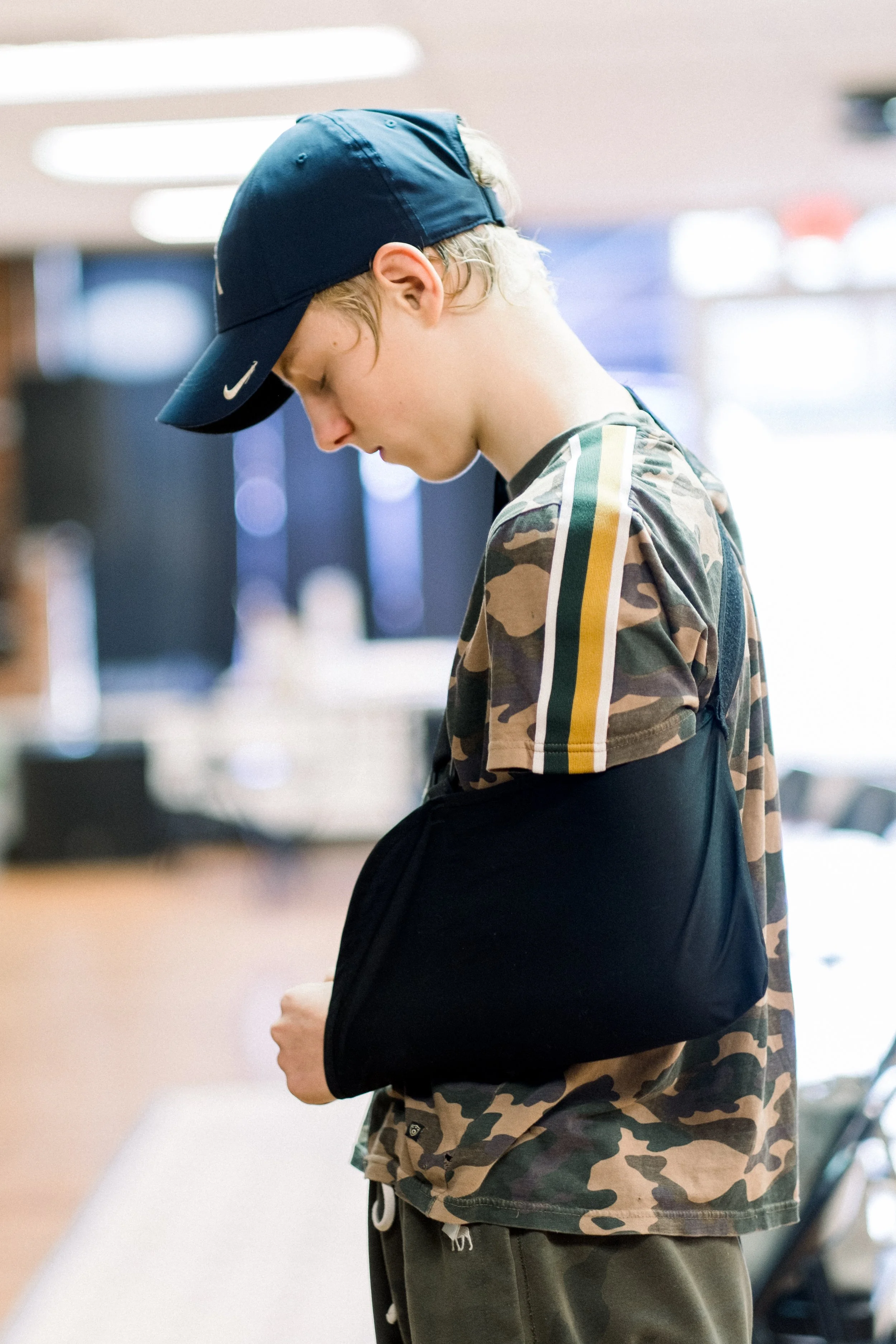 A young person wearing a blue Nike cap, camouflage t-shirt with yellow and green stripes on the sleeves, and carrying a black backpack, standing indoors.
