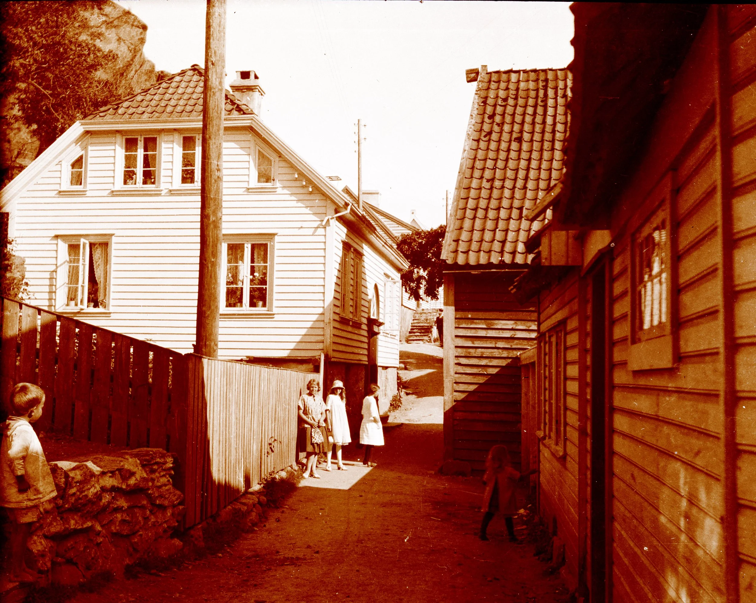 Gammel svart-hvitt fotografi av en gate med hus og barn som står i en rekke, omgitt av gjerder og trær.