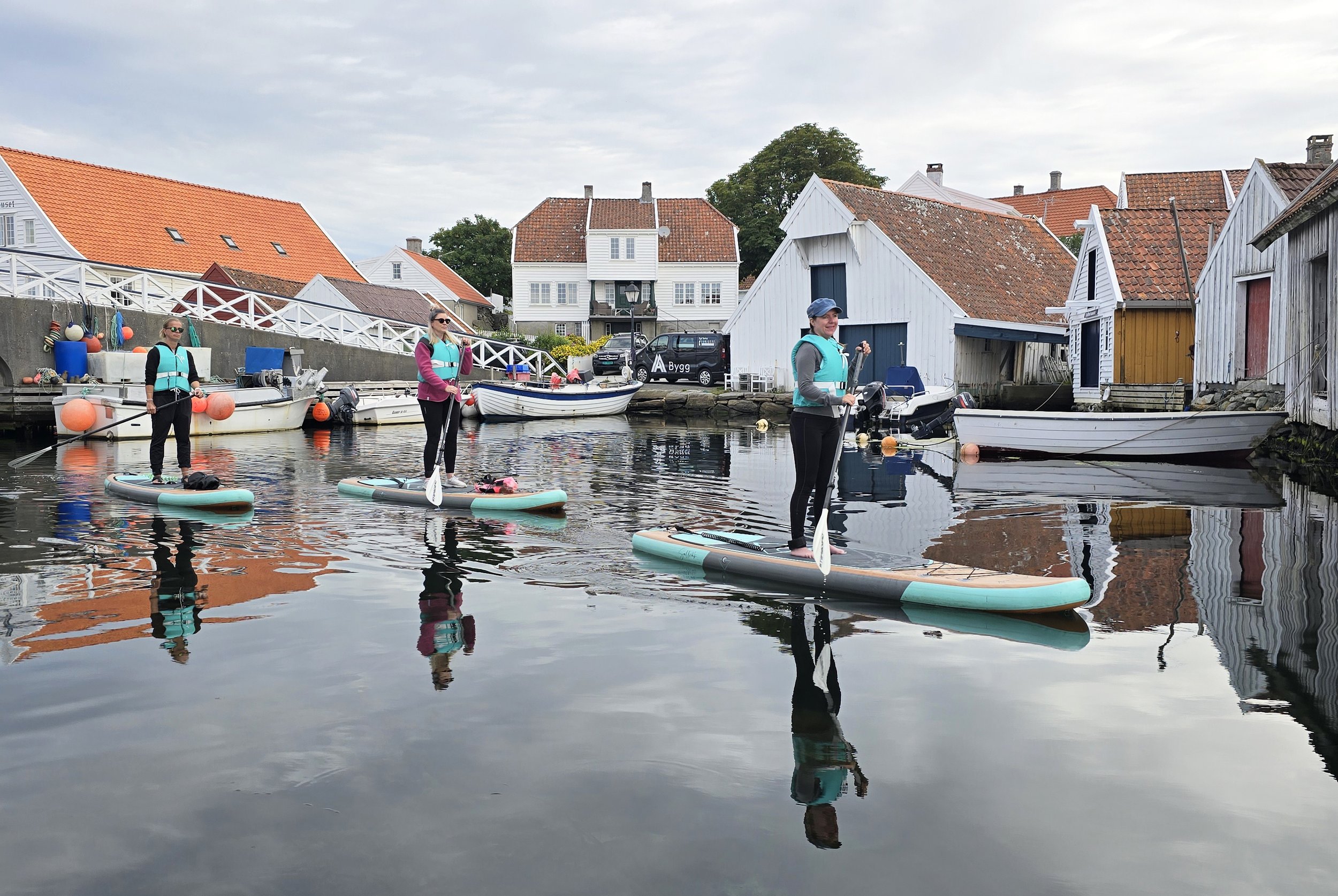 Tre personer som står på paddleboarder i vannet nær en havn med små hus med røde og hvite tak i Norge.
