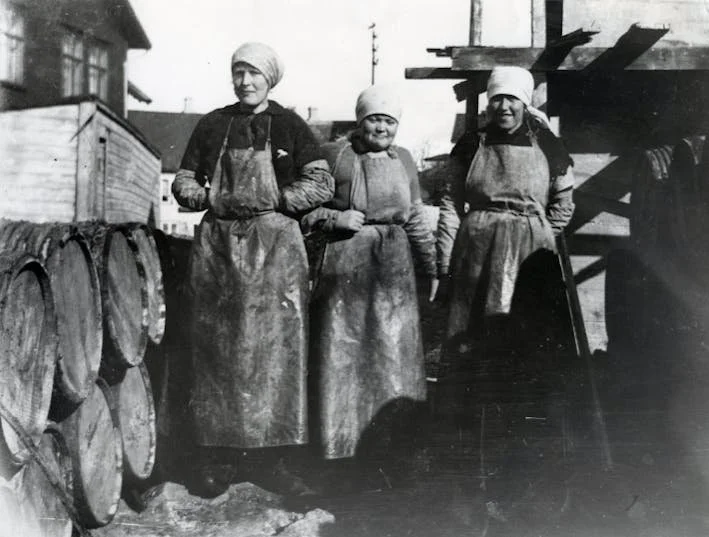 Tre kvinner i gamle klær og forkle på en utendørs arbeidsplass, omringet av tømmerstokker og annet tømmer, sannsynligvis fra tidlig 1900-tall.