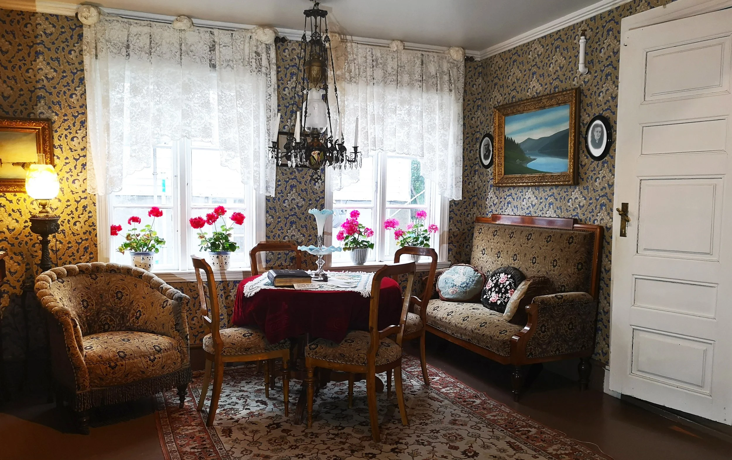 Et rom med møbler, blomster og kunst på veggene, lyse gardiner foran vinduene, og en taklampe i midten.