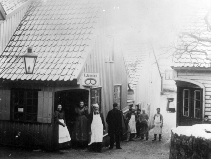 En gruppe mennesker utenfor en trebygning, noen av dem i arbeidsklær, i en gammel bygate. Svart-hvitt foto, trolig fra tidlig 1900-tall.