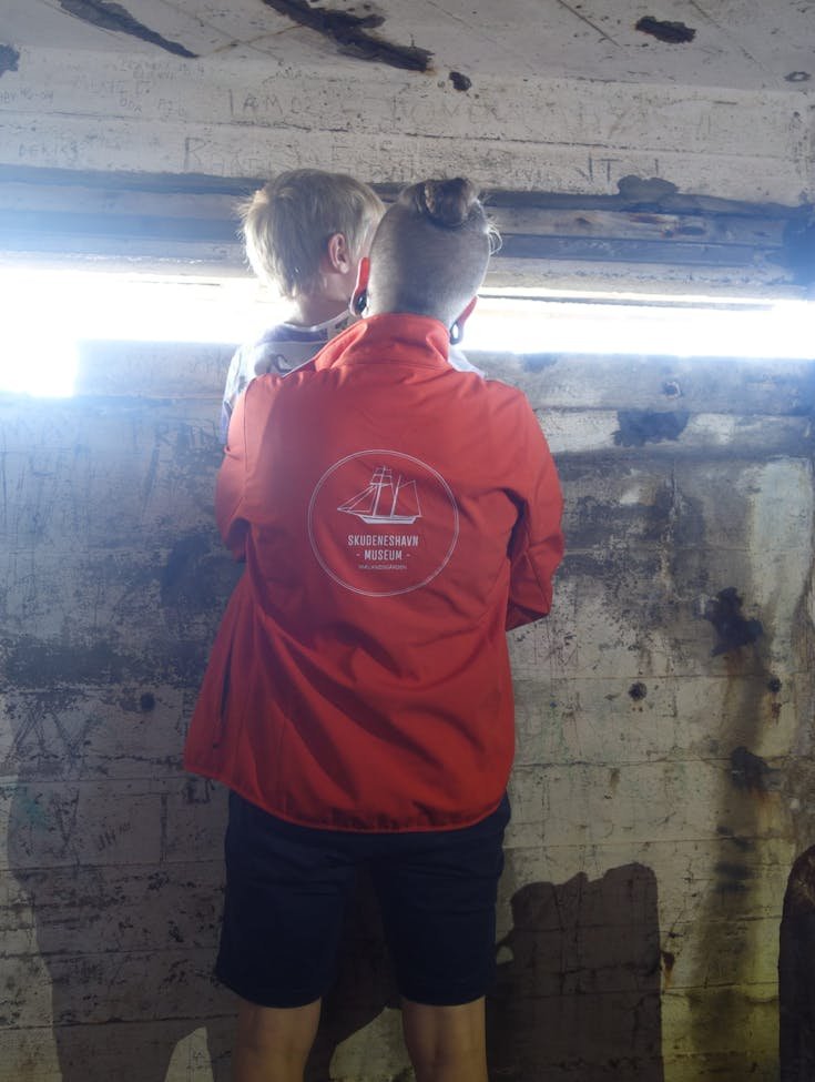 To personer står i et mørkt rom med en smal stripe med lys foran dem, en av dem har på seg en rød jakke med logoen til Skudeneshamn Museum på ryggen.