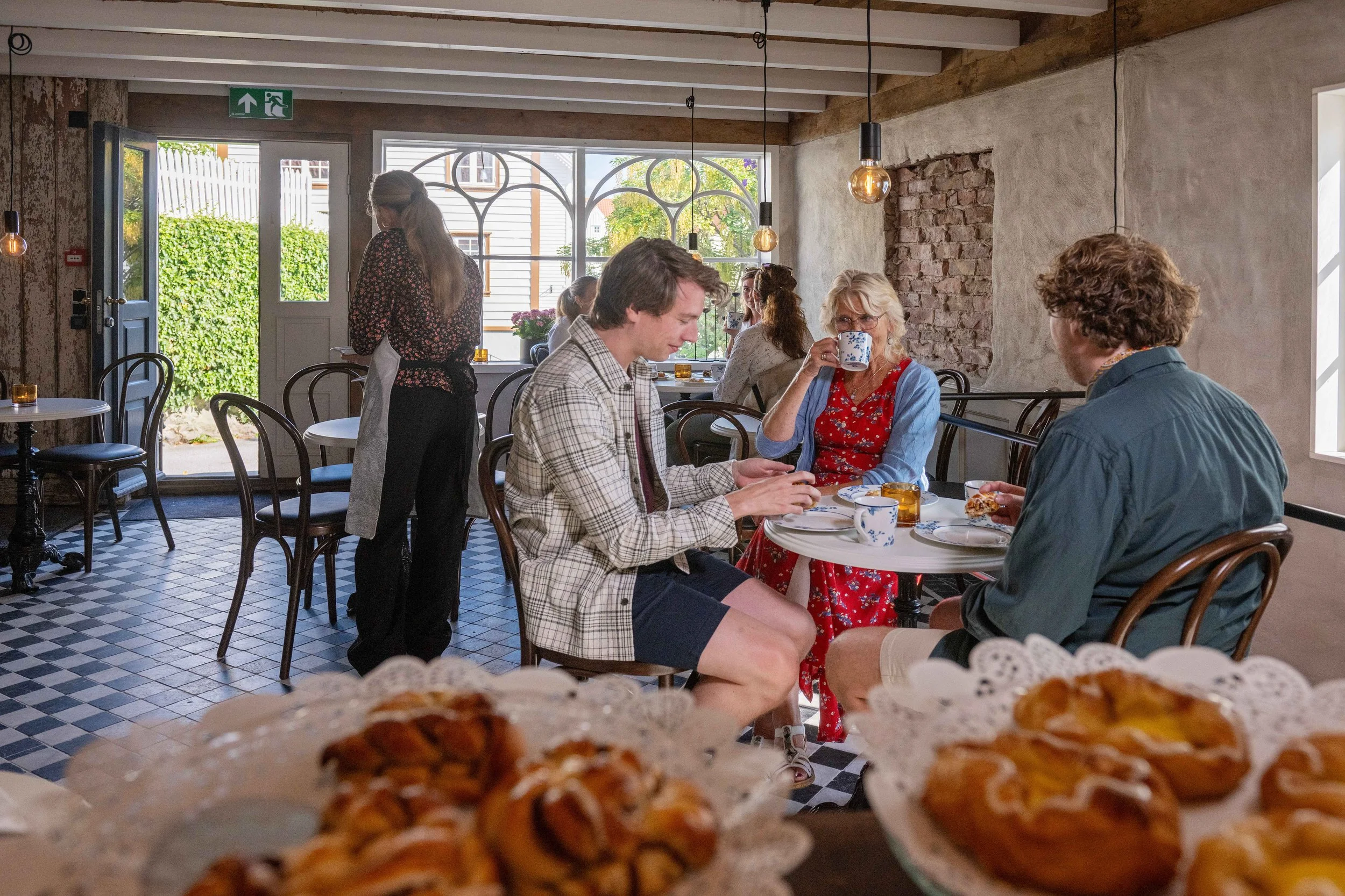 Innendørs kafe med mennesker som spiser og drikker, grønne planter utenfor vinduet, og bakverk i forgrunnen.