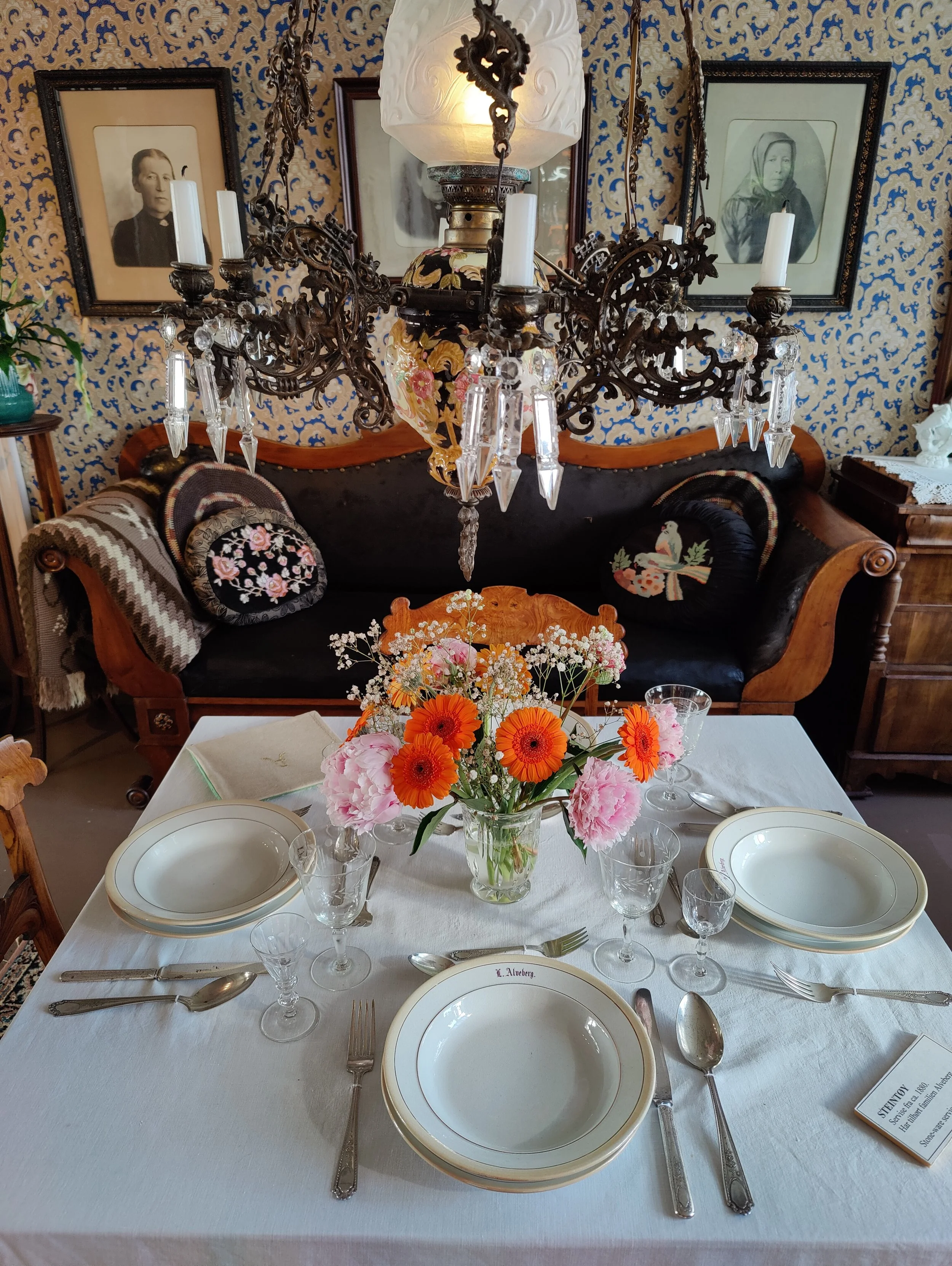 Et bord dekket for et måltid med hvit duk, to tomme serveringsfat, bestikk, vinglass og vannglass. Midt på bordet står en vase med blomster. Bak det er en sofa med puter og en stor lysekrone henger over bordet, med bilder på veggen bak.