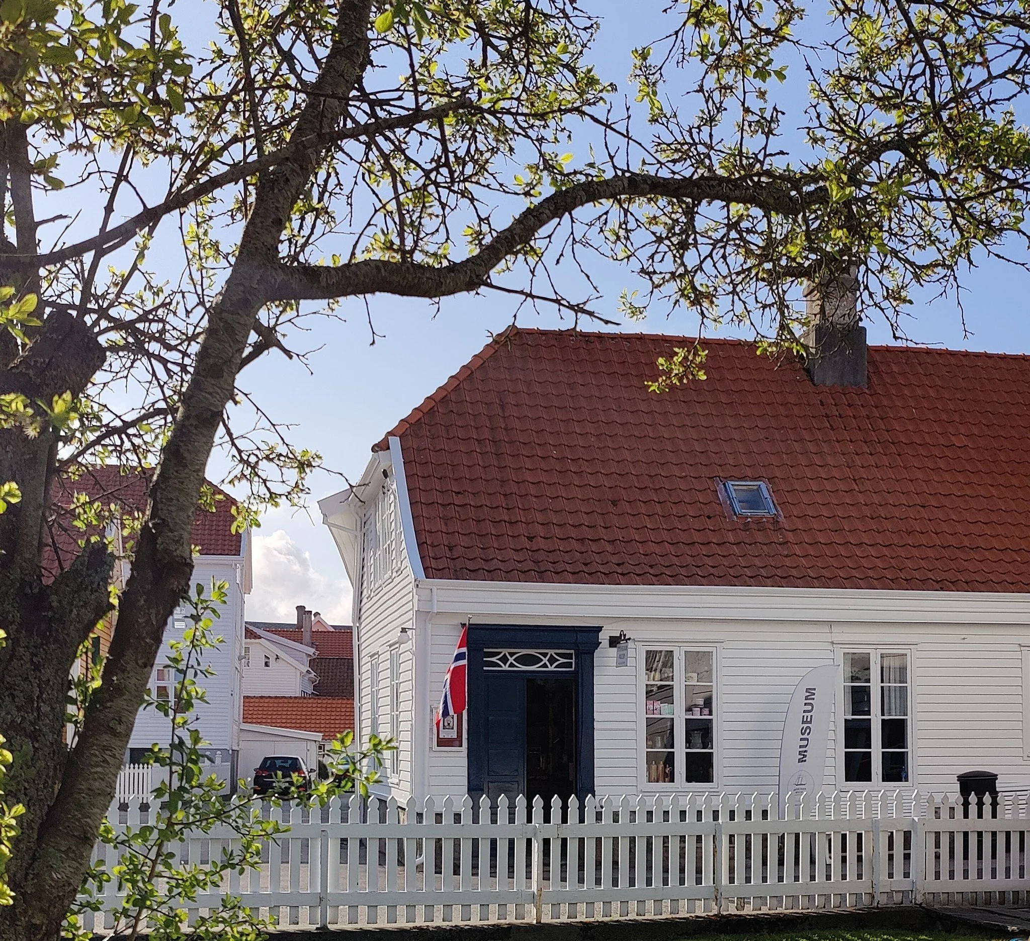 Et hvitt hus med svart dør og store vinduer, omgitt av hvit hekk, med en norsk flagg foran. En stor trebauttre og en rødfarget tak med en liten takluke kan sees.