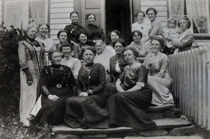 Gruppe av kvinner og menn foran en hytte, uskarpt svart-hvitt bilde.