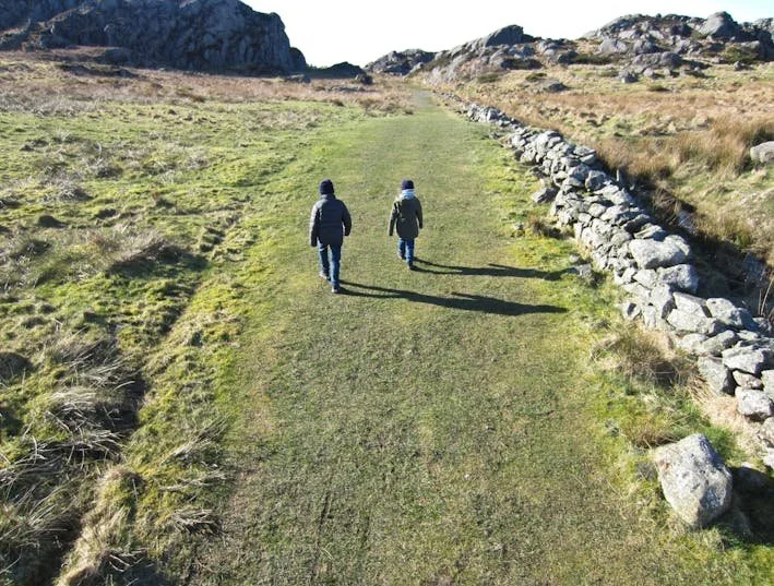 To personer går på en gruslagt sti i et åpent landskap, omgitt av gress og fjell, med en steinmur på høyre side.