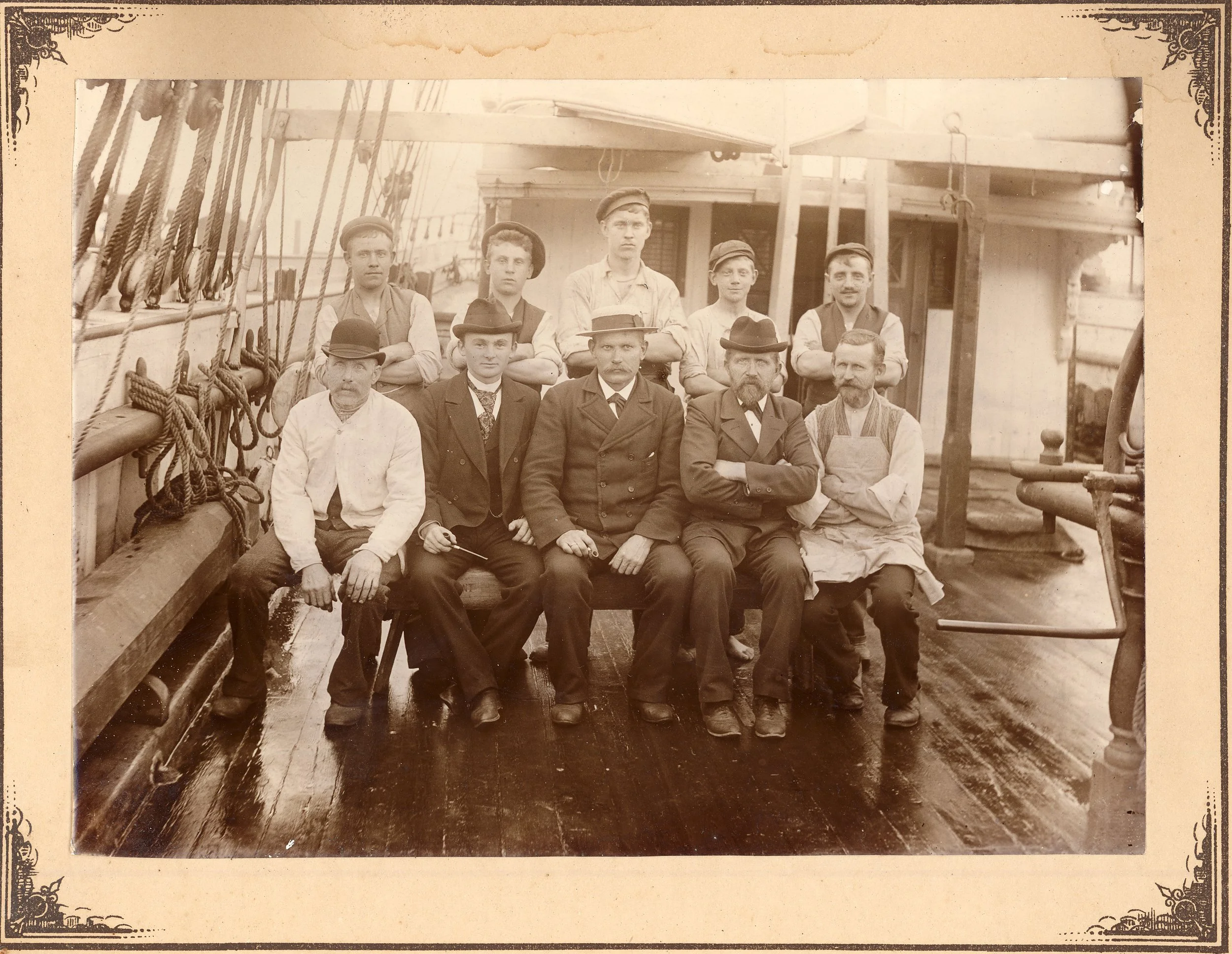 Sepiafarget gruppebilde av ni voksne menn, noen med hatter, på en trebrygge eller dekk, muligens på en gammel båt eller skipsdekk.
