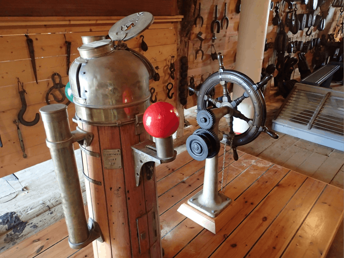 Vintage maskin med roterende hjul og koblinger, plassert på en trebenk i et arbeidsrom med verktøy på vegg.