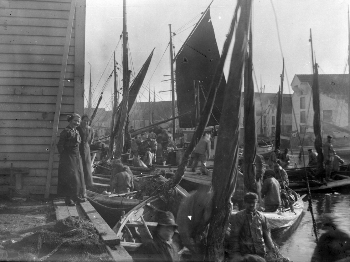 Bilde av en havn med båter og mennesker, flere båter med seil er fortøyd ved kai, og folk står og sitter rundt båtene.