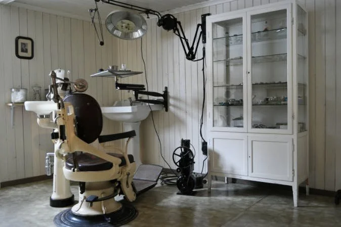 Et gammelt tannlegekontor med tannlegeutstyr, tannlegekrakk, og et oppbevaringsskap med glassdører.