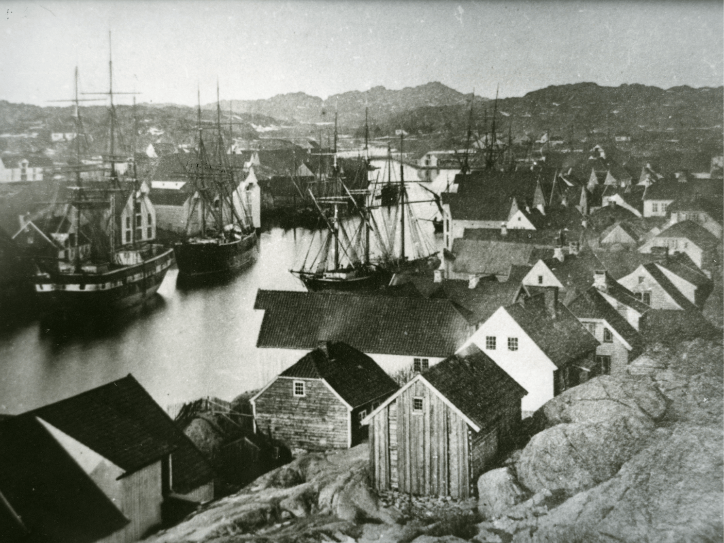 Historisk svart-hvitt bilde av en liten sjøby med tre båter i havnen og små hus langs kysten med fjell i bakgrunnen.