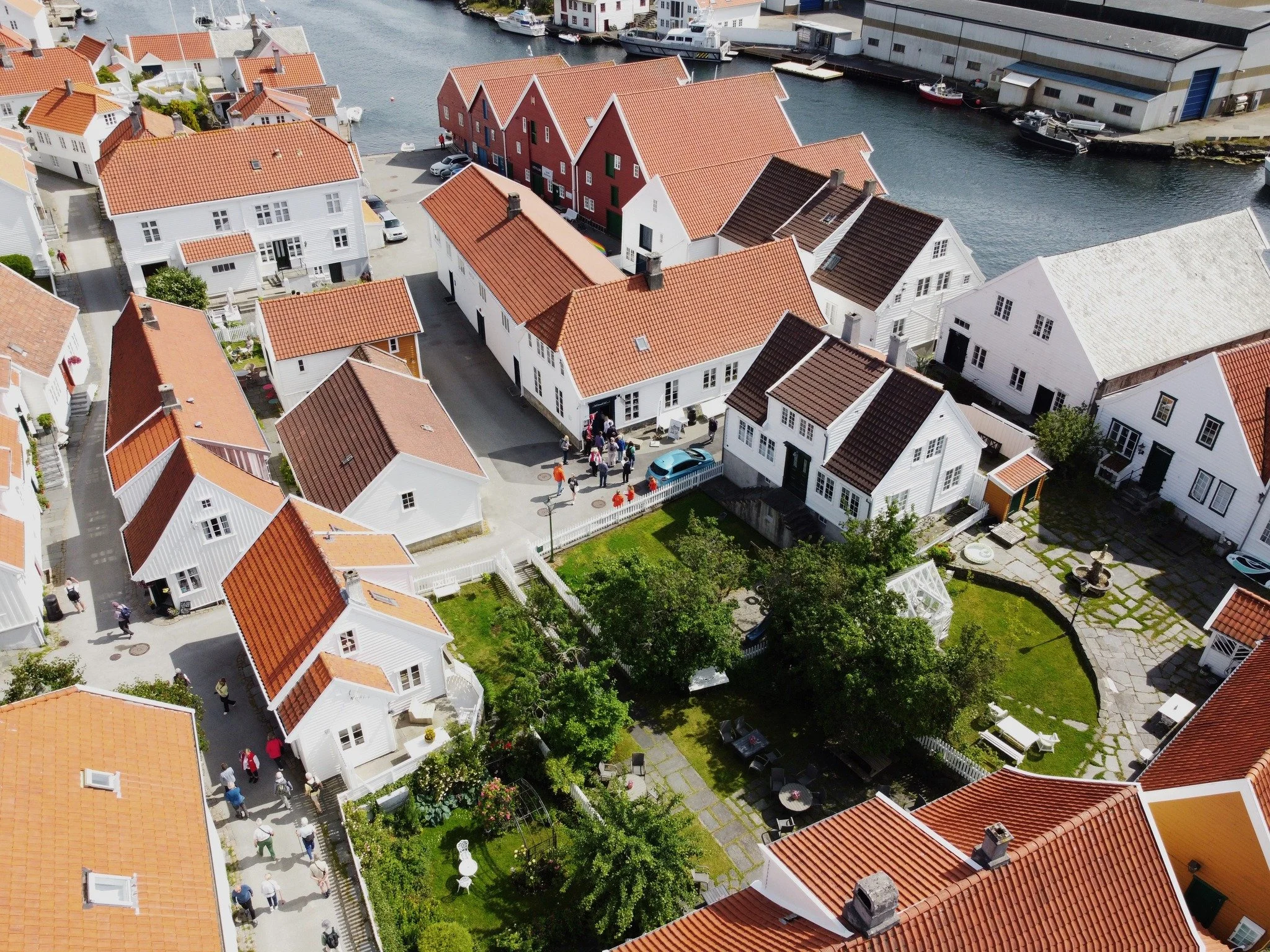 Luftfoto av en liten kystby med hvite hus med røde tak nær vannet, mange folk går og står i gatene, og det er båtmerker og en havn i bakgrunnen.