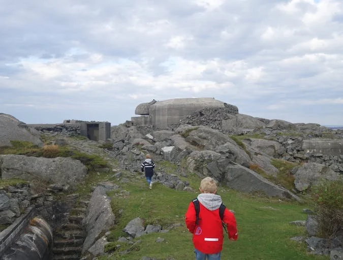 To barn går oppover en sti mot en stor, gammel, forfallen bunkers i et landskap med store steiner og gress, under en overskyet himmel.