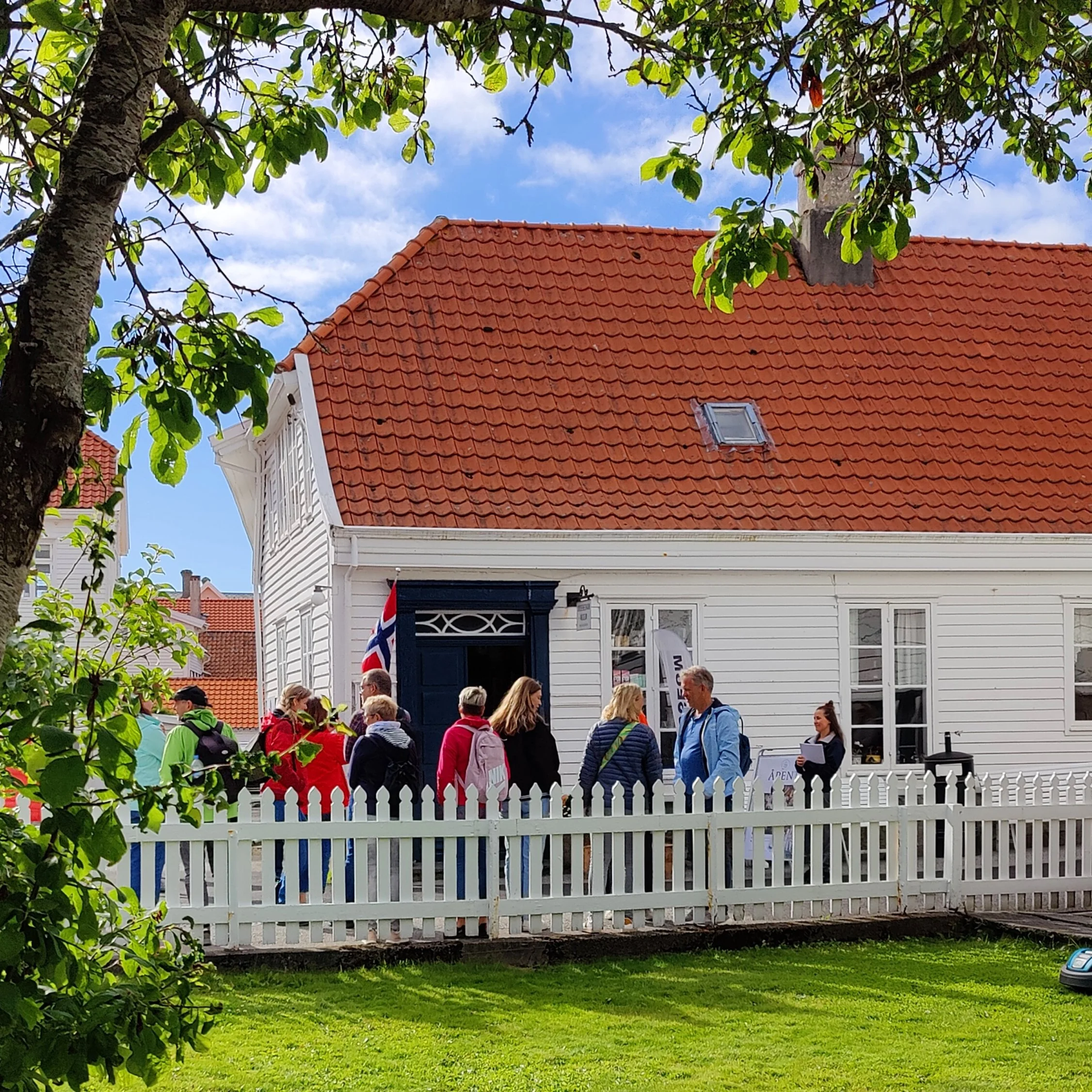 Et hvitt hus med rødt tak og en hvit hekk foran, omgitt av grønne trær og folk som står på gaten.