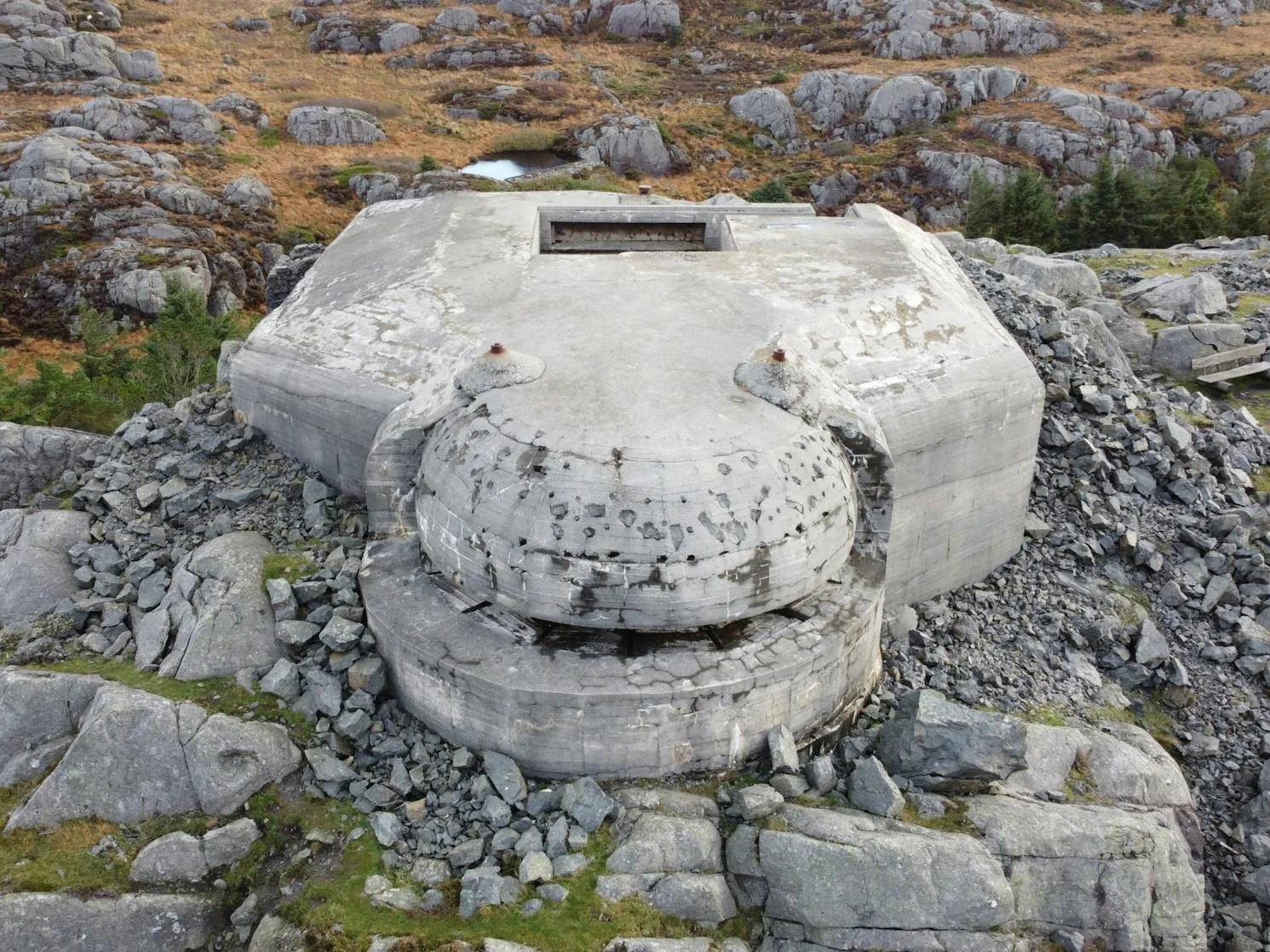 En solid, rund betongstruktur plassert i en berglandskap, omringet av steiner og fjell, med noe vegetasjon i bakgrunnen.