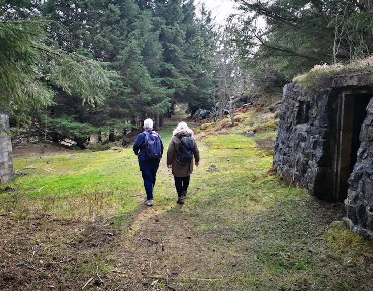 To personer går along en skogssti i et grønt område med trær og stabilt terreng, med bygninger i stein på høyre side.