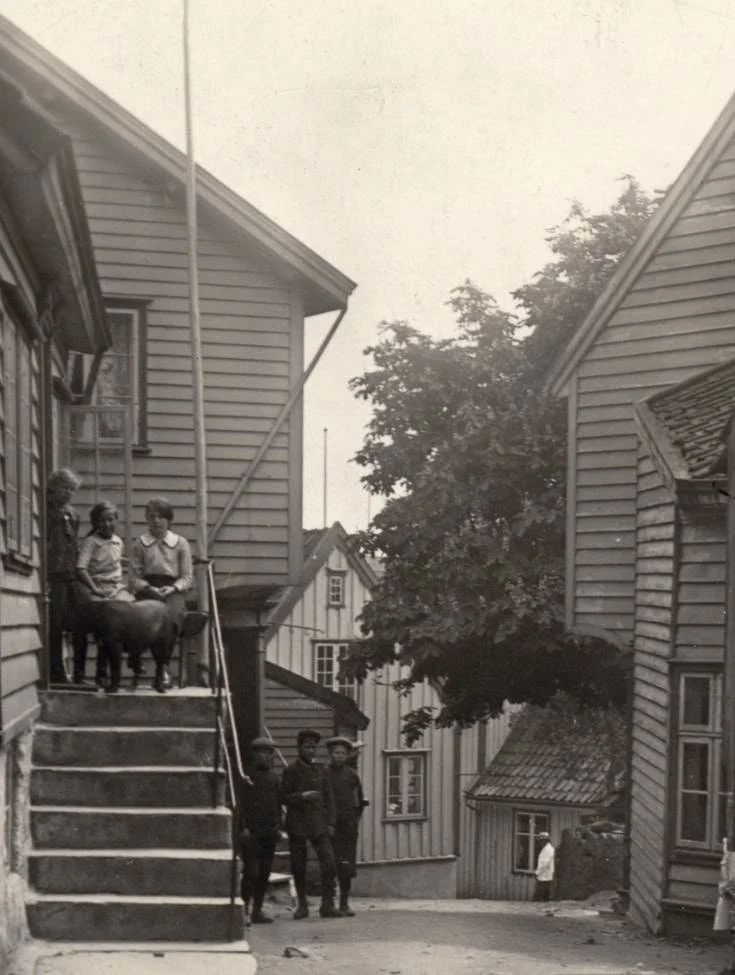 Sølvgrå bygninger med mennesker, noen på trappene og noen på gaten, en hund og et tre.