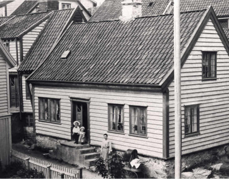 Et svart-hvitt bilde av en enkel trehus med tre vinduer og en dør, med tre personer foran inngangen, og en hage med gjerde.