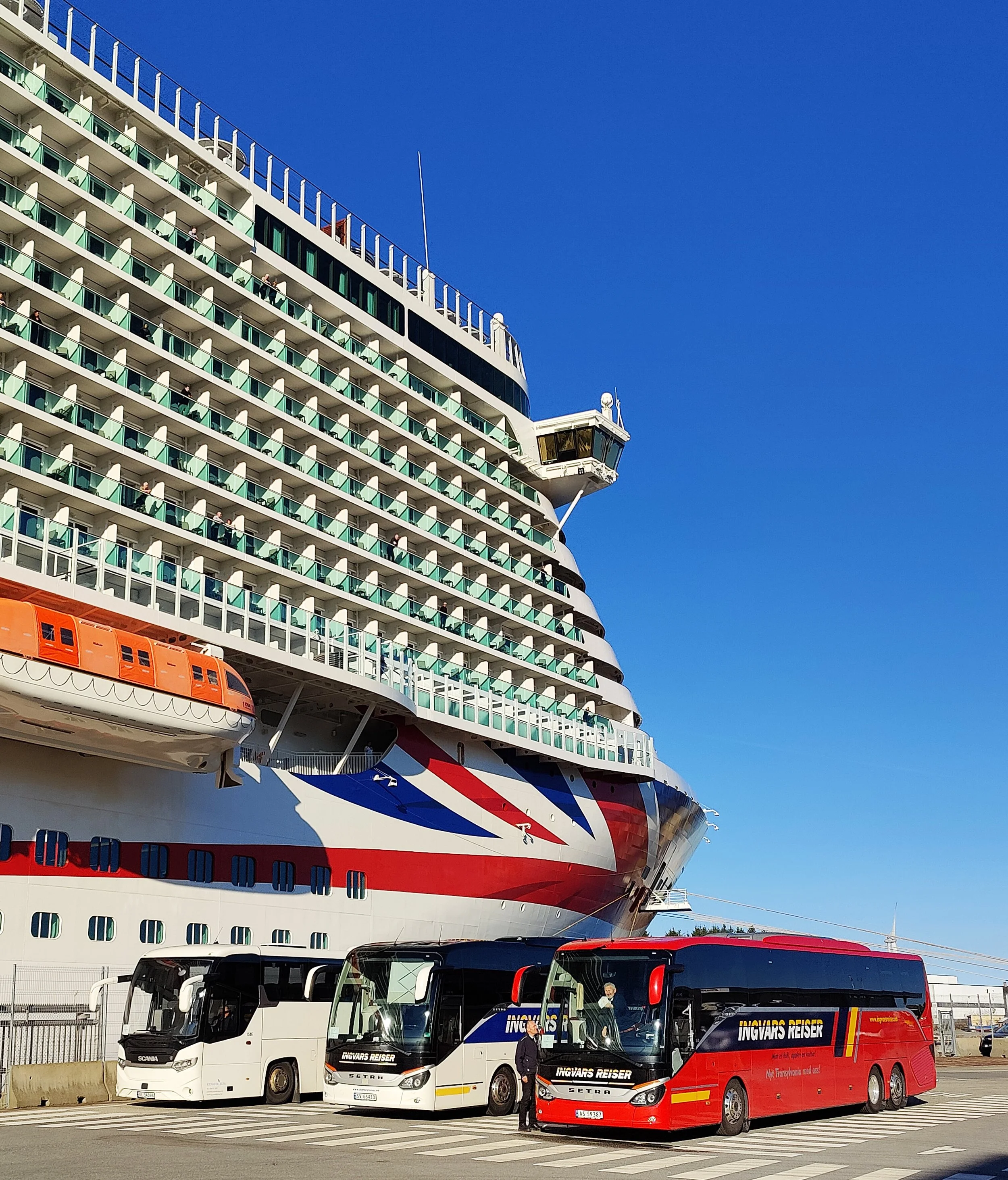En stor cruiseflytende med flere dekk og store vinduer. Innvendig kan man se passasjerer og utstyr. I forgrunnen er det en buspark med tre busser i ulike farger og en person som står ved den røde bussen. Under et vindu på cruiseskipet er det et orange livbåt.