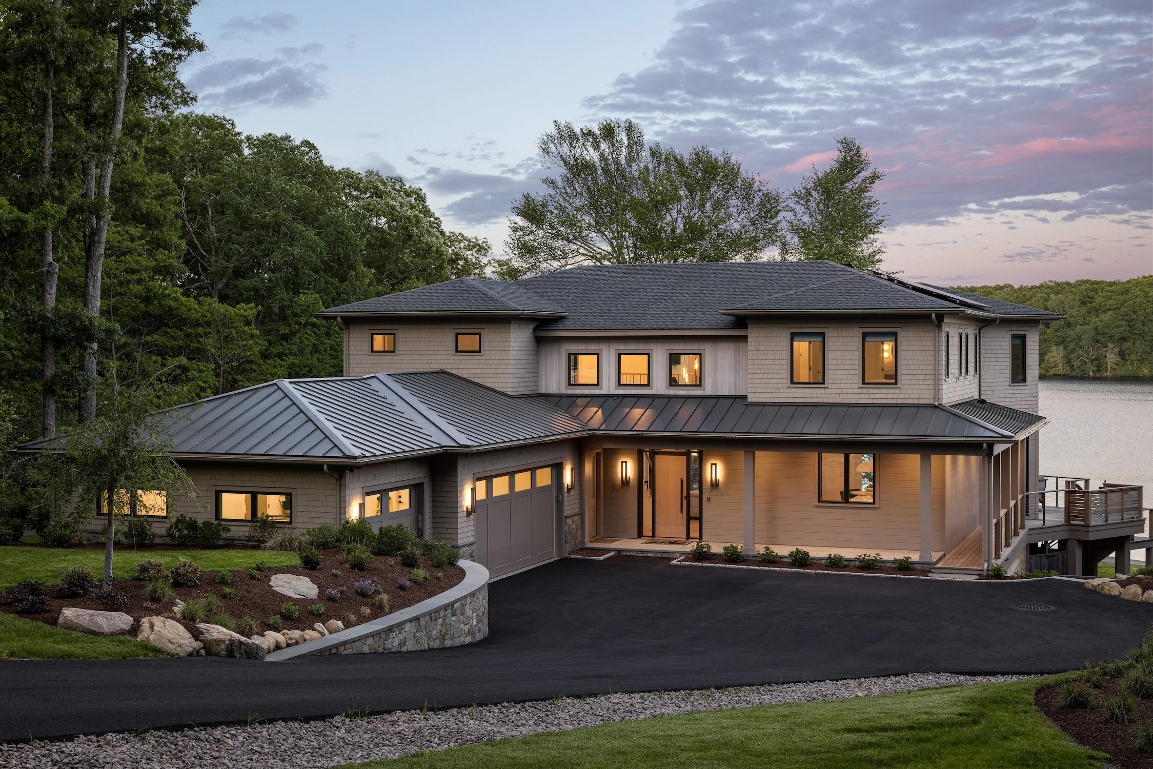 A modern two-story house with large windows, situated near a body of water, surrounded by lush greenery, with a sunset sky in the background.