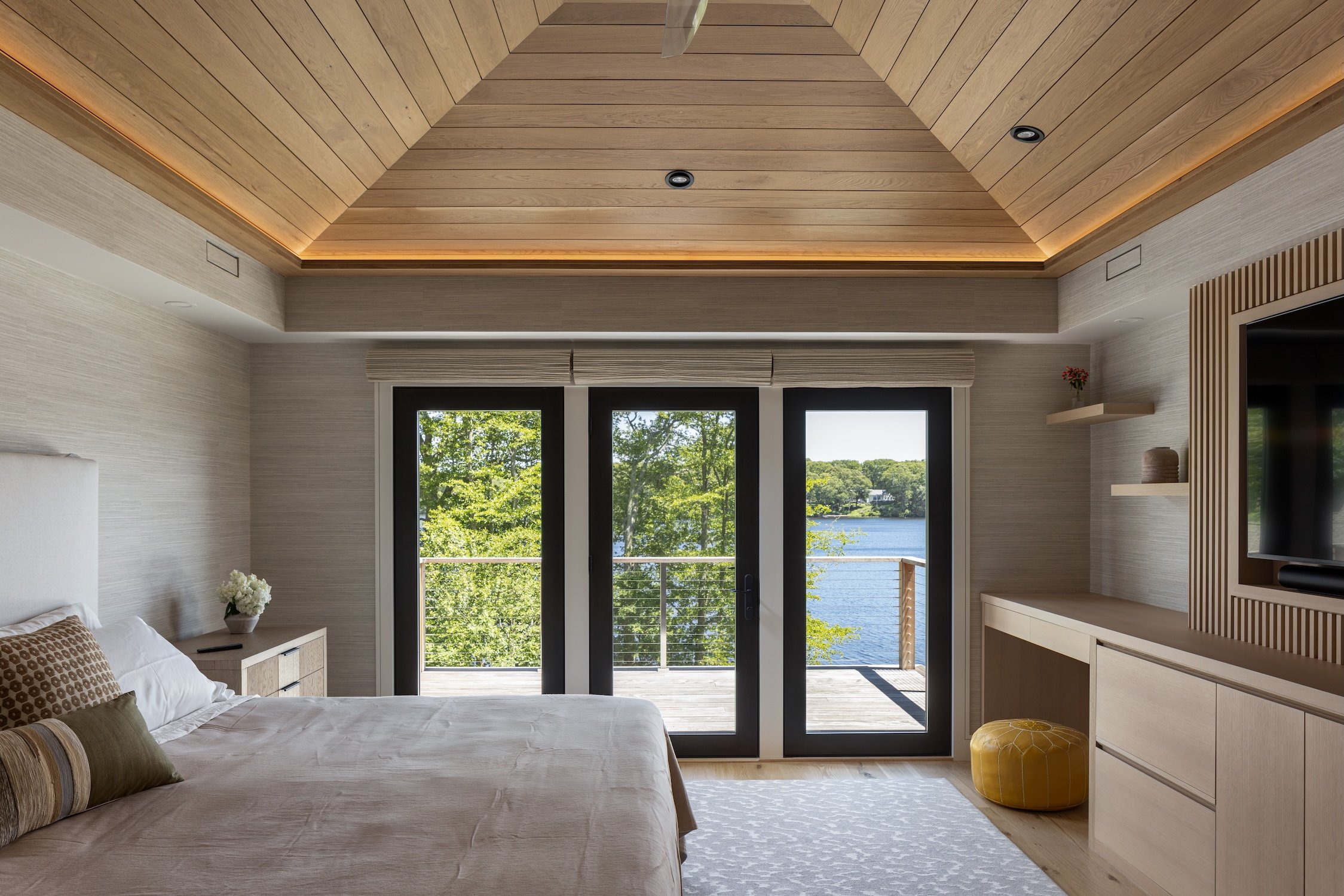 Modern primary bedroom with a vaulted white oak ceiling, custom millwork, recessed lighting, and large glass doors overlooking the water.