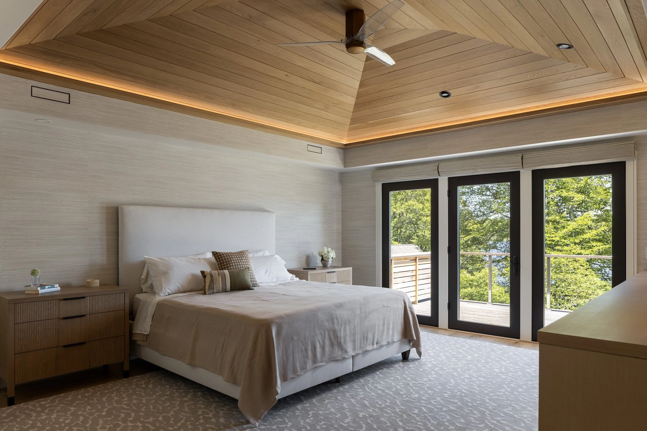 Modern primary bedroom with a vaulted white oak ceiling, custom millwork, recessed lighting, and large glass doors overlooking the water.