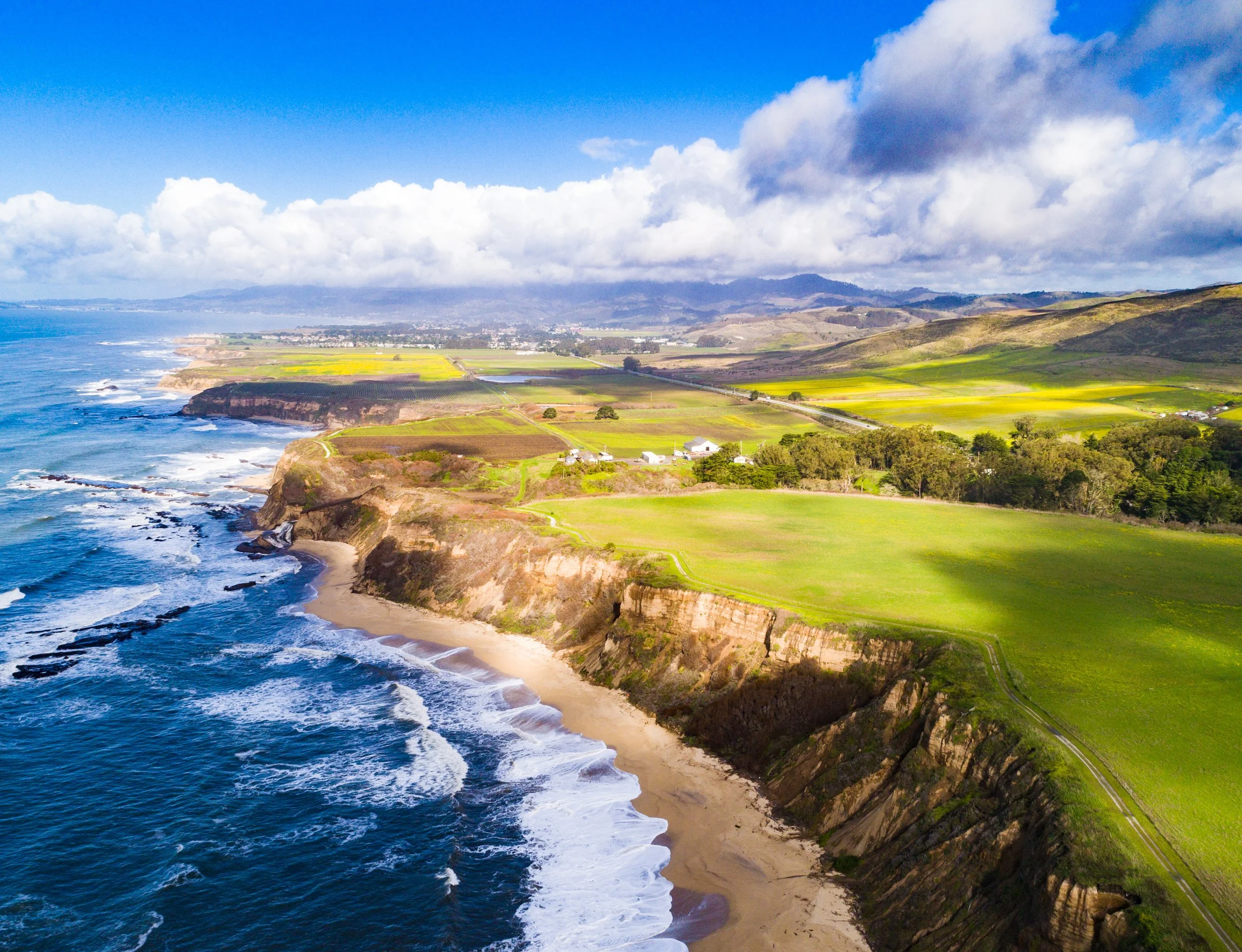 Coastal landscape with cliffs, sandy beach, green fields, and blue ocean under partly cloudy sky.