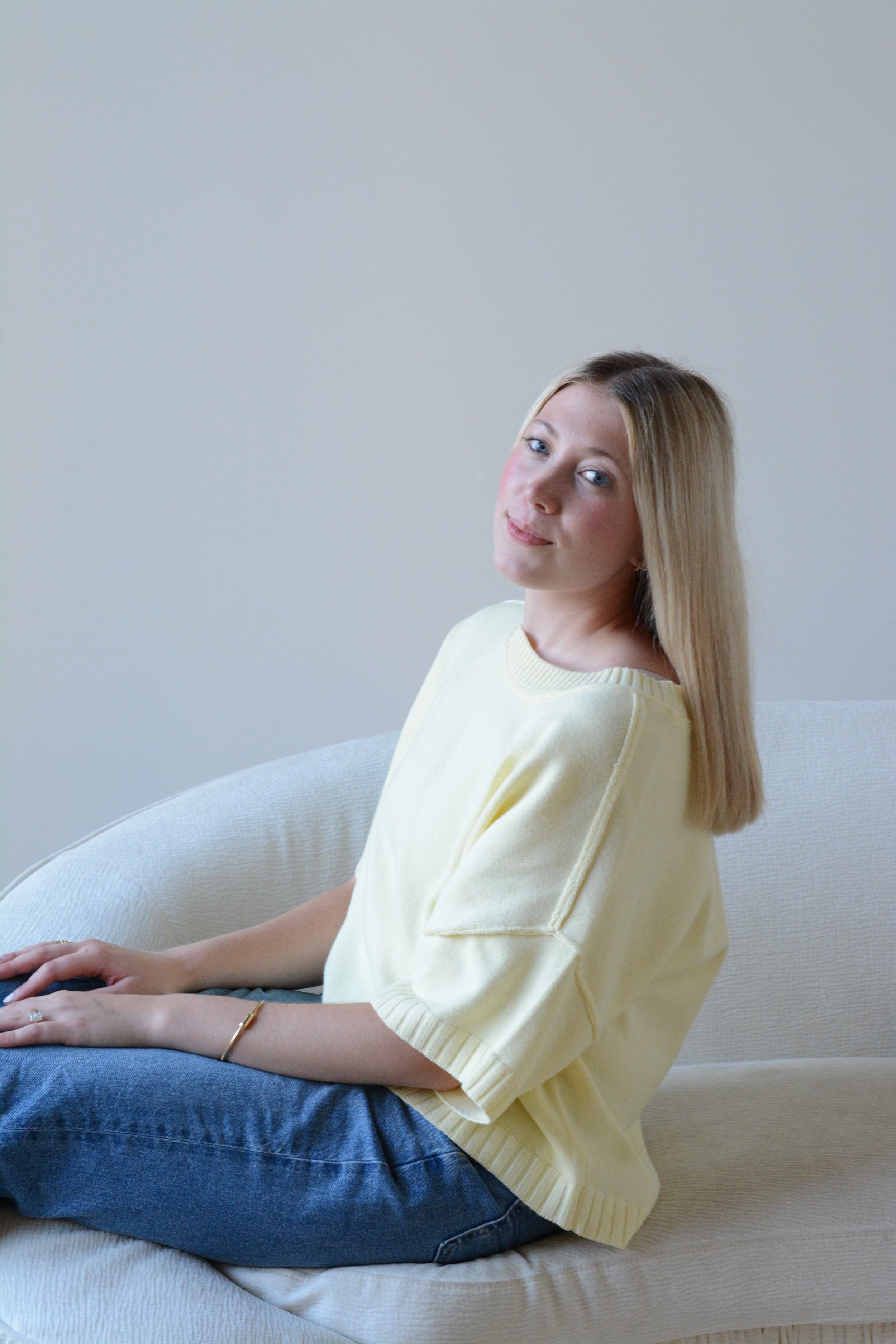 A young woman with blonde hair, wearing a light yellow shirt and blue jeans, sitting on a light-colored sofa against a plain wall, looking at the camera with a slight smile.
