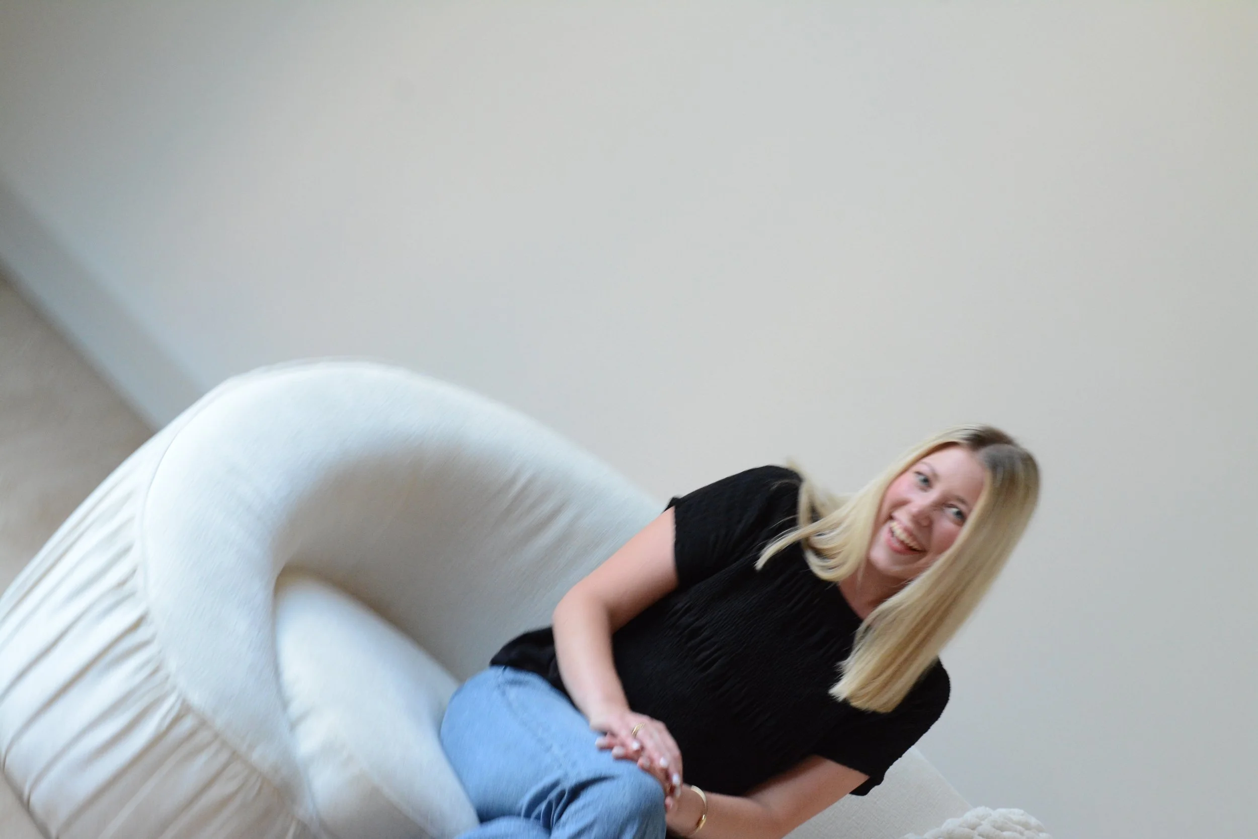 A woman with blonde hair wearing a black top and blue jeans, sitting on a cream-colored sofa, smiling at the camera in a room with a plain white wall.