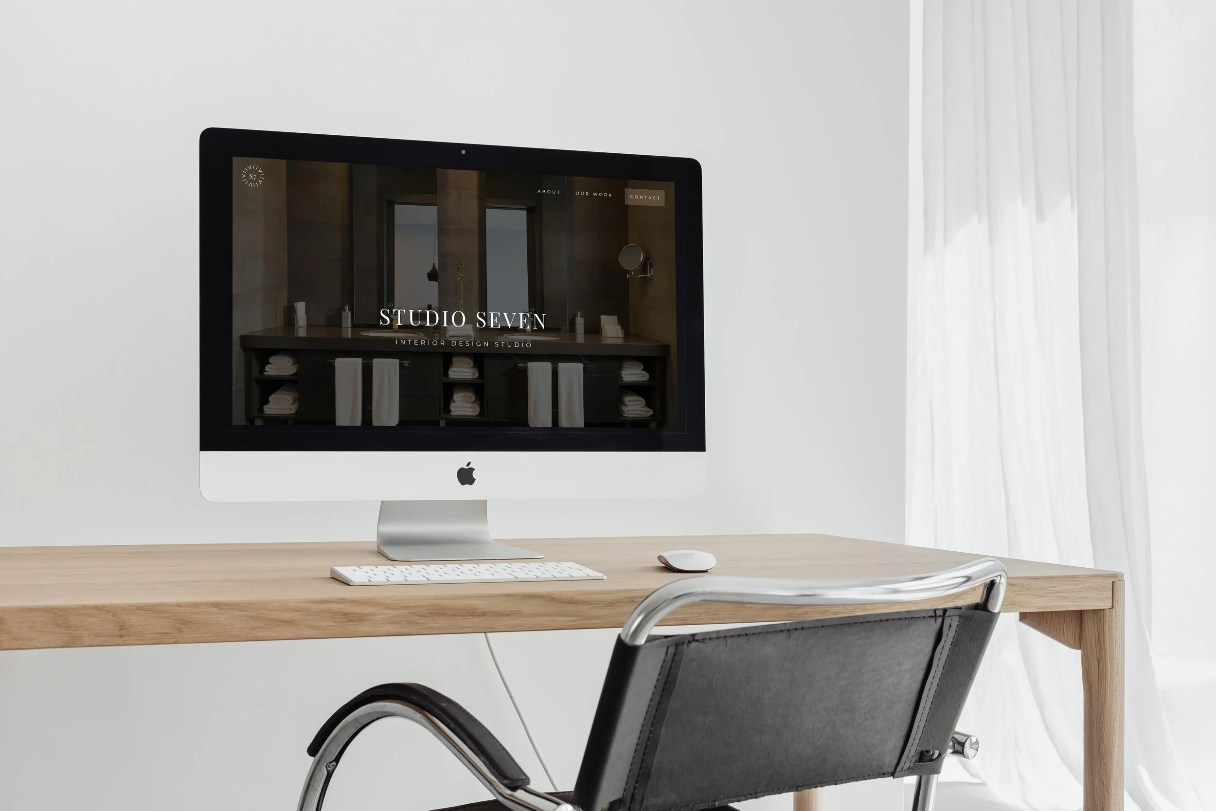 An Apple iMac computer with a keyboard and mouse on a light wood desk, displaying a website for 'Studio Seven Interior Design Studio,' with a minimalist room with white walls and curtain in the background.