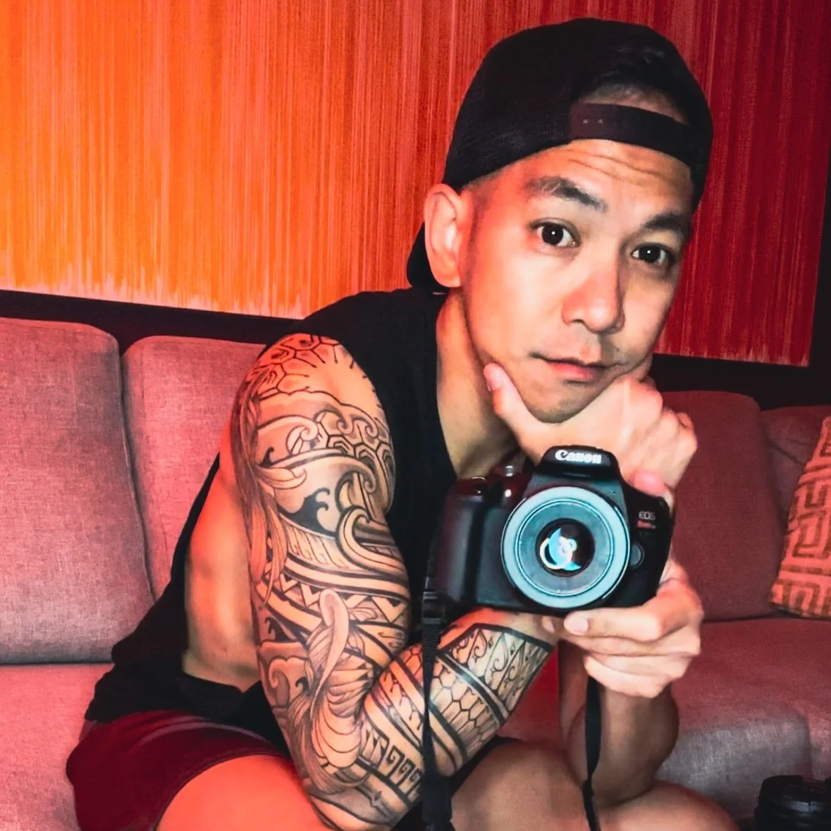 Wilford with tattoos on his arm holding a Canon camera, sitting on a couch with an orange and red modern painting in the background.