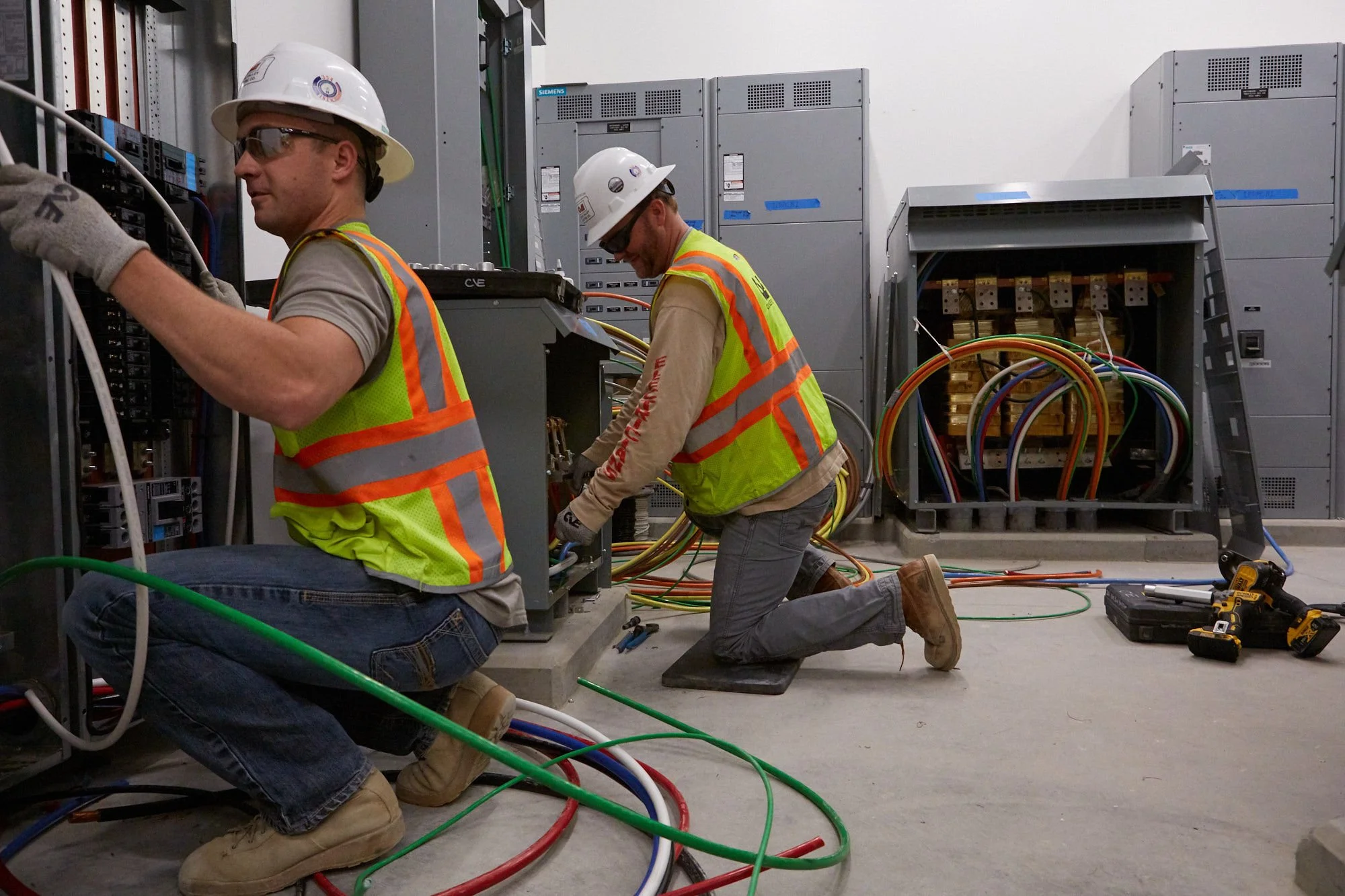 Two electricians working on electrical panels in an industrial setting, wearing safety helmets, vests, and gloves, with tools and colorful wires around them.
