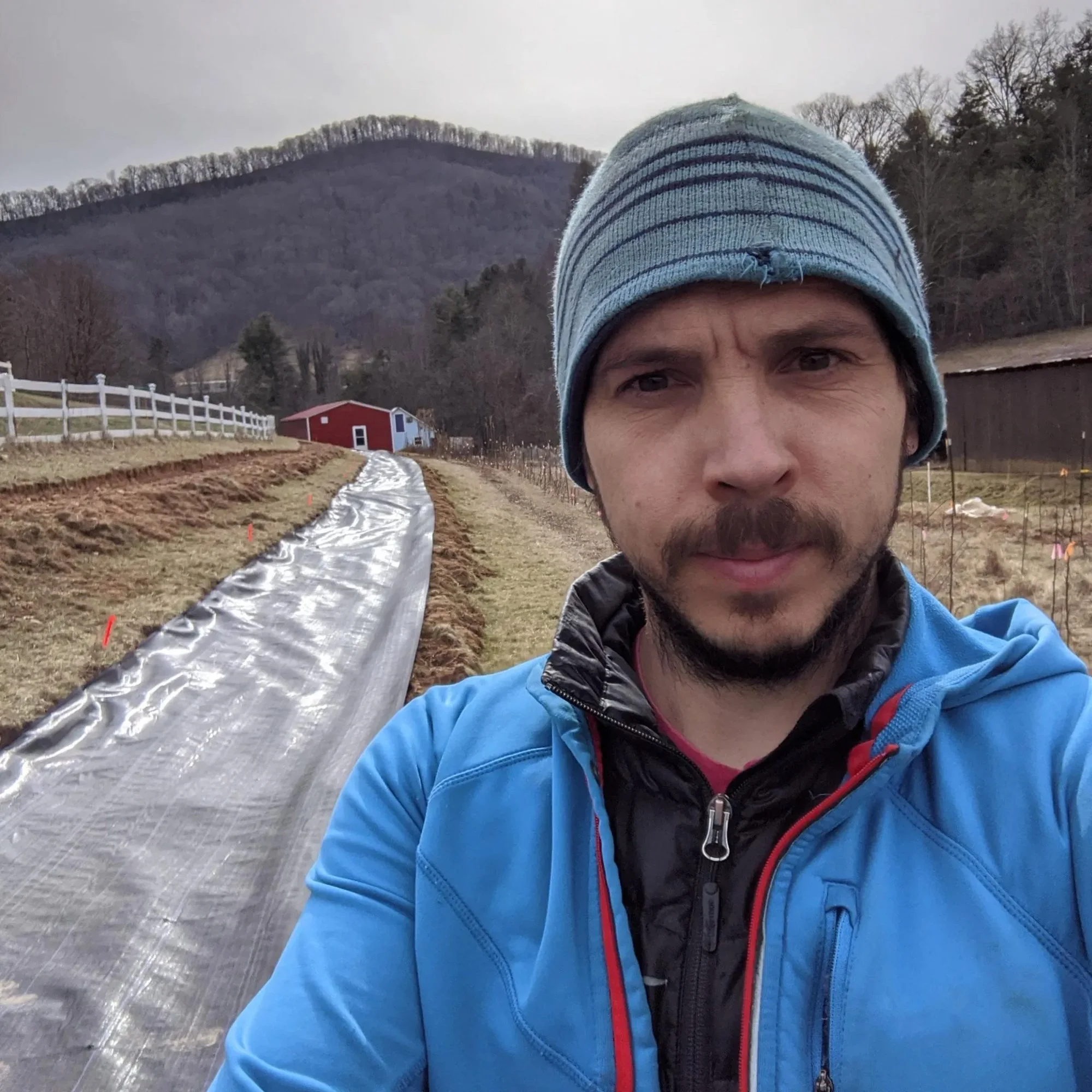 A man in a blue jacket and striped beanie taking a selfie outdoors on a farm or garden with a plastic-covered garden bed, a red and white building, a wooden fence, and mountains in the background.