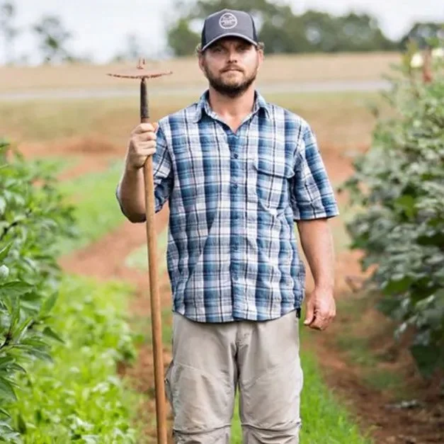 A man stands in an outdoor farm or garden, holding a rake, wearing a plaid shirt, beige pants, and a baseball cap, with greenery and rows of plants in the background.