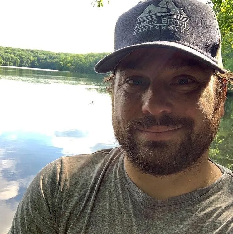 A man with a beard and mustache, wearing a gray t-shirt and a navy blue cap with a logo that reads 'AMES BROOK CAMP GROUND,' smiling at the camera near a peaceful lake with trees in the background.