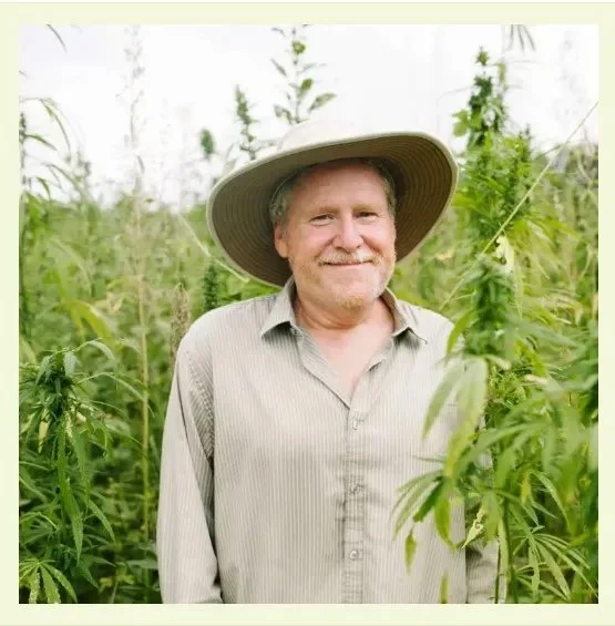 A man with gray hair and a beard smiling, standing among tall cannabis plants in a greenhouse, wearing a wide-brimmed straw hat and a light-colored button-up shirt.
